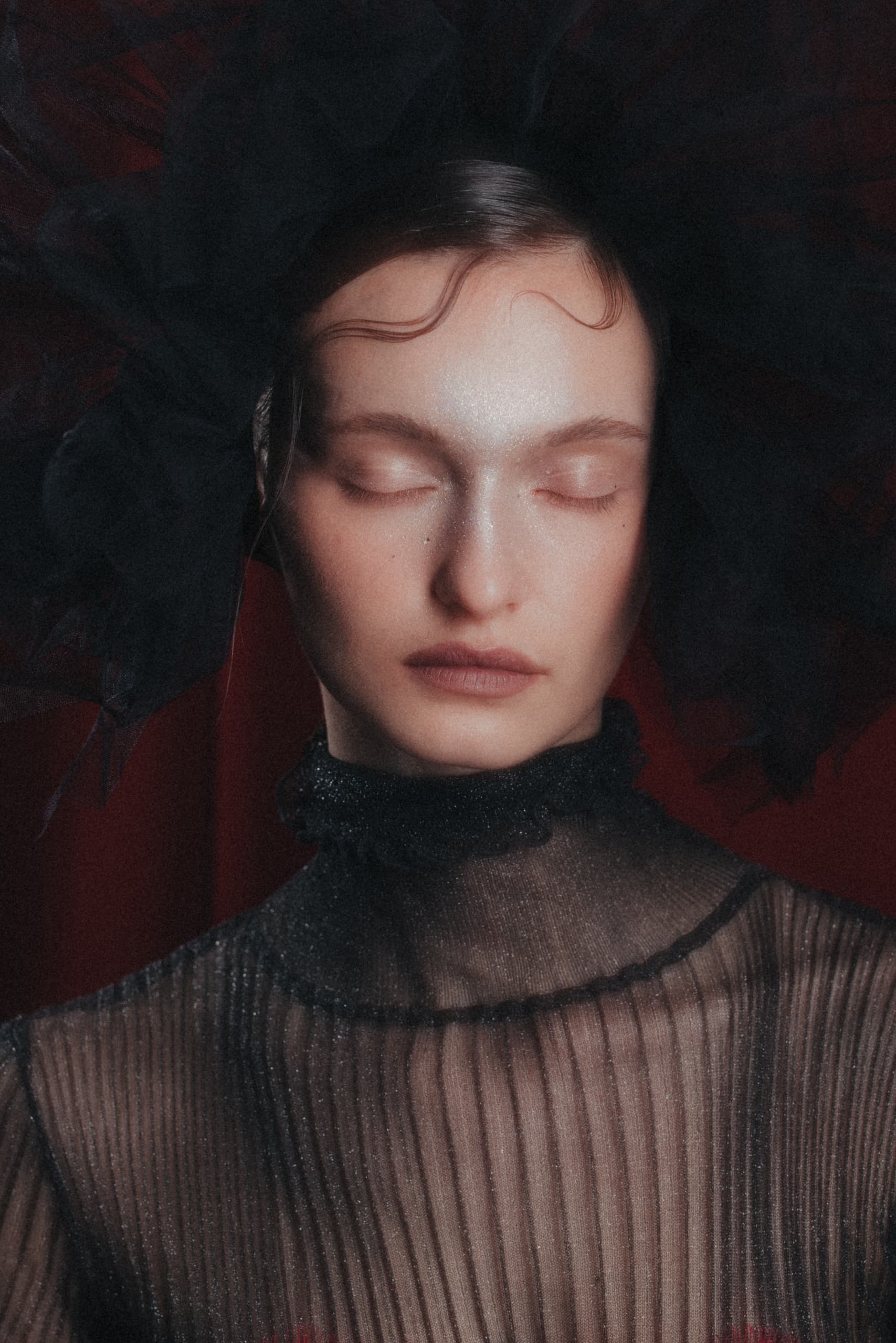 Woman in sheer black dress with dark headpiece, eyes closed, dramatic fashion look.