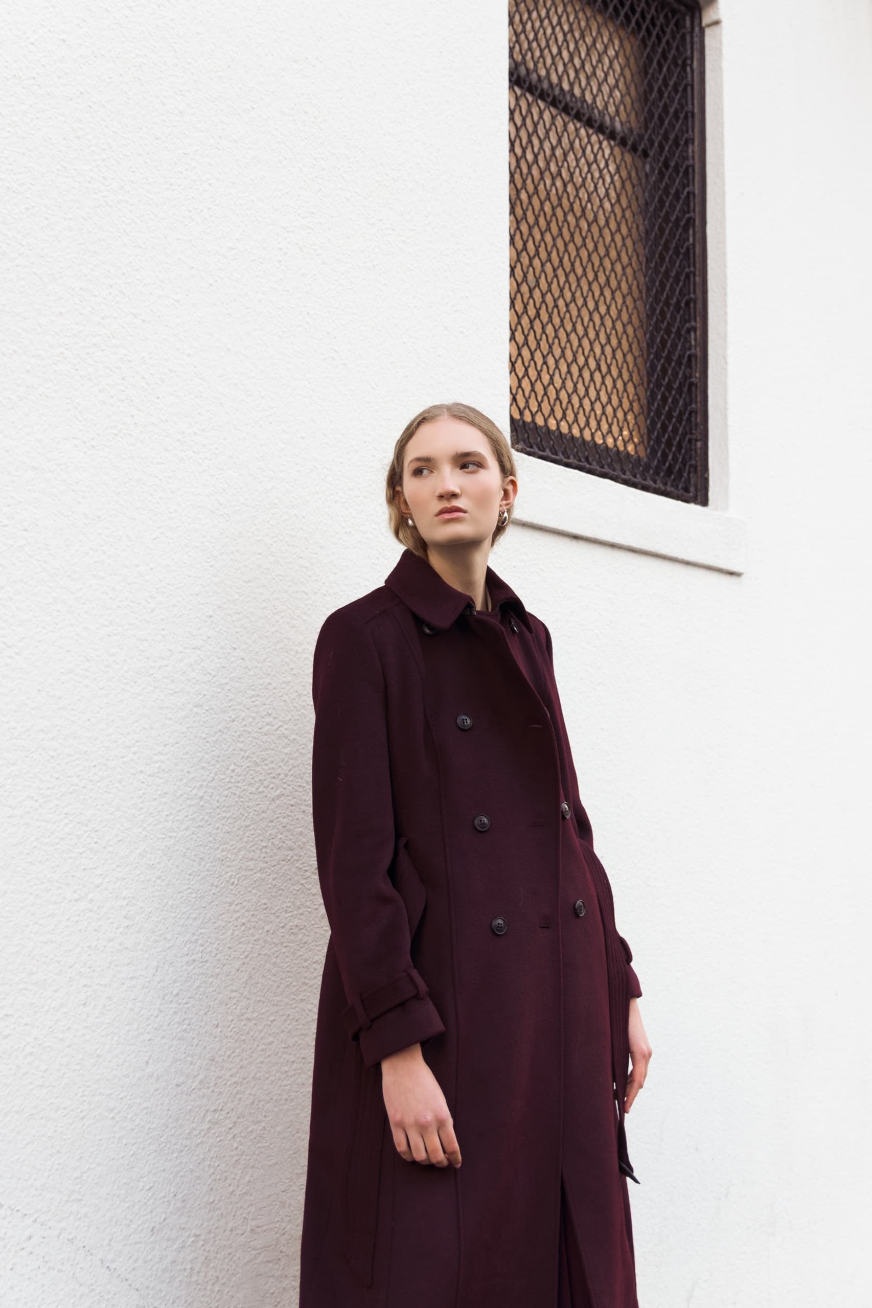 Fashionable woman in burgundy coat against white wall background