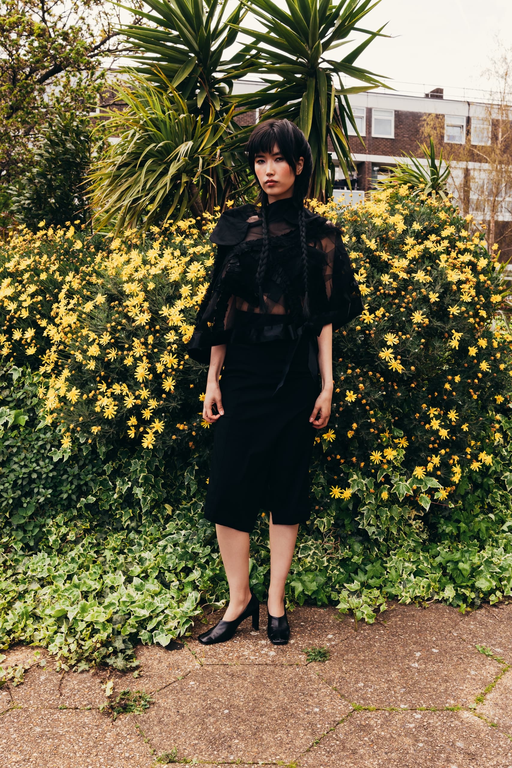 Woman in black dress stands by yellow flowers and green foliage.