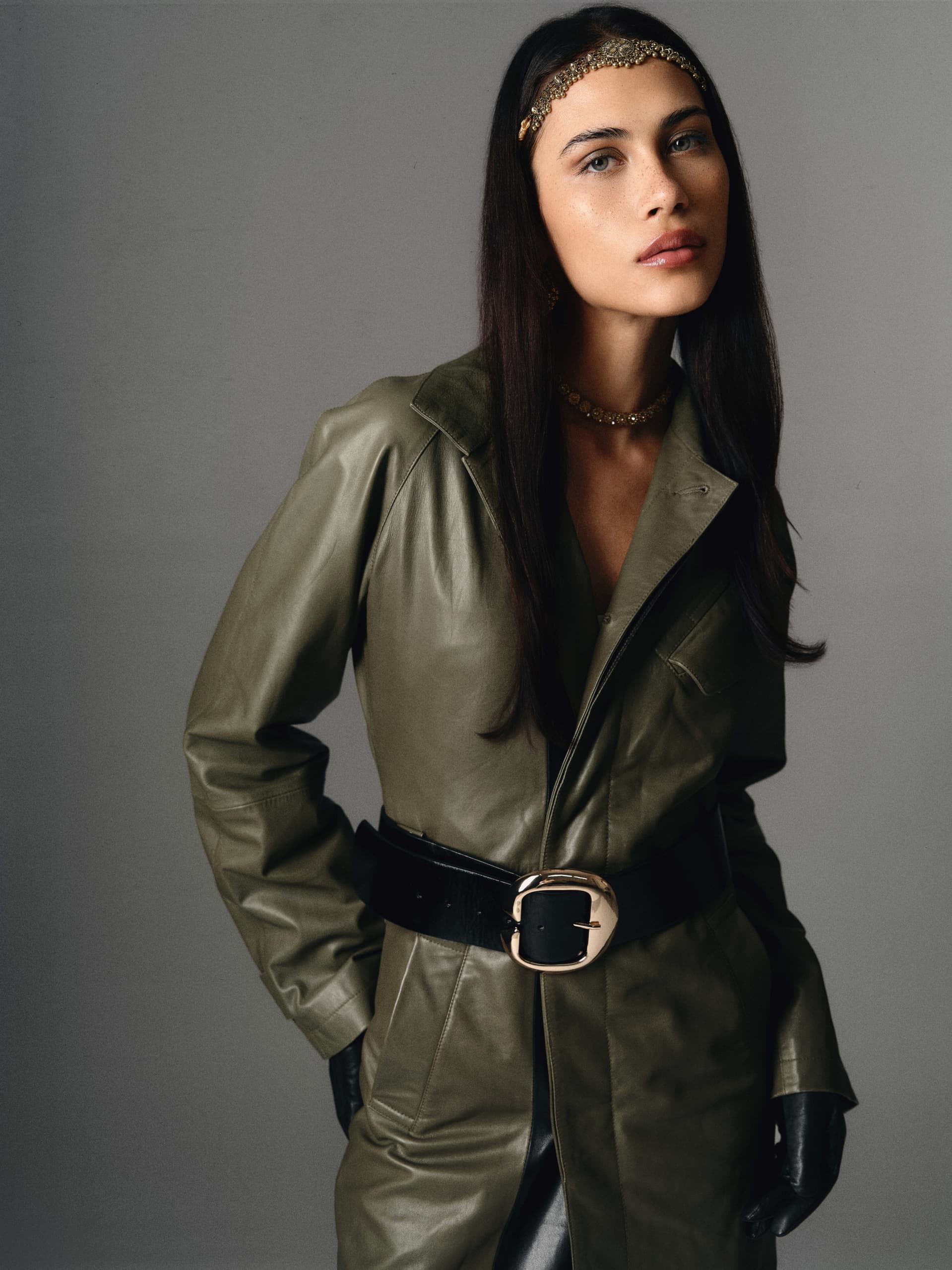 Fashion model in green leather coat and gold accessories against a gray background.