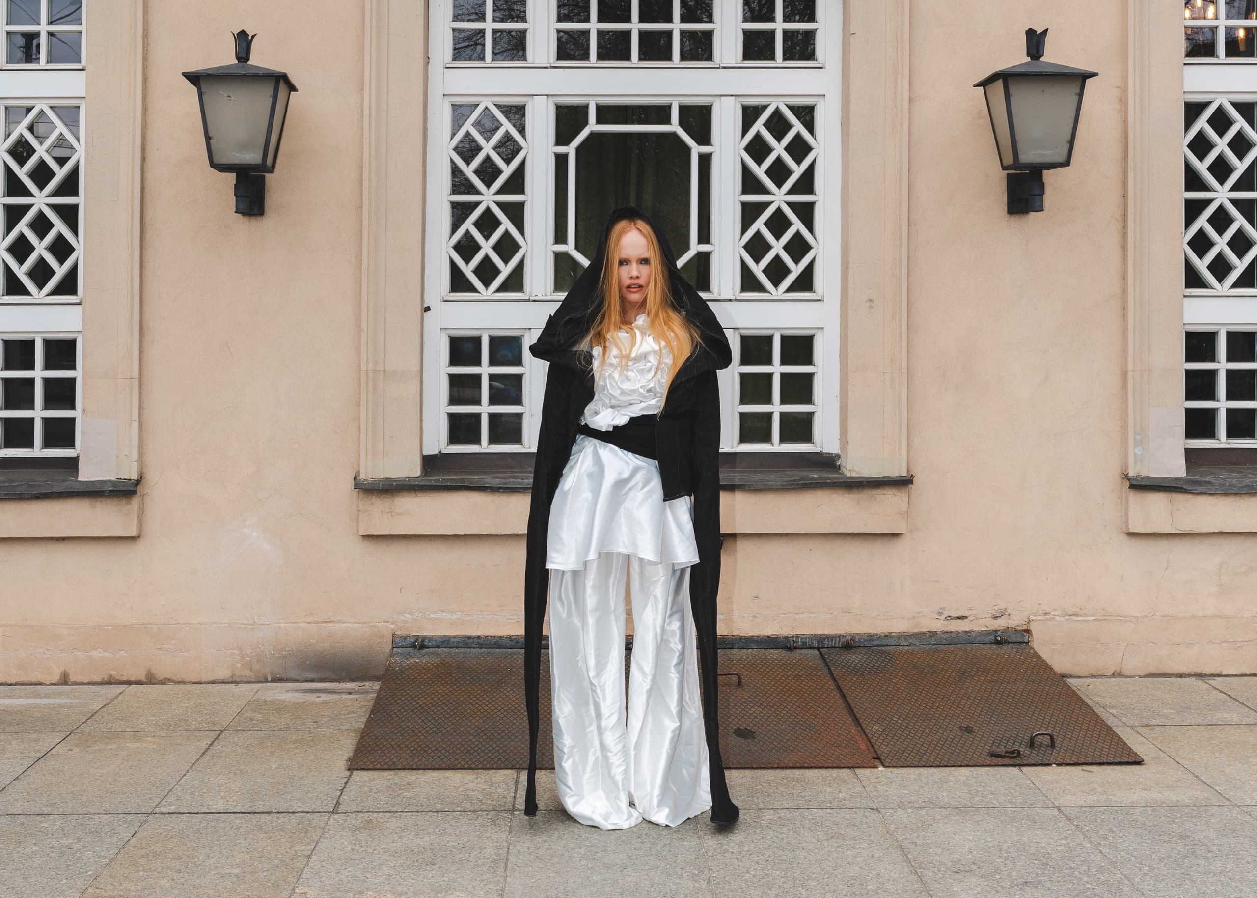 Fashionable woman in black and white outfit stands before elegant windows.