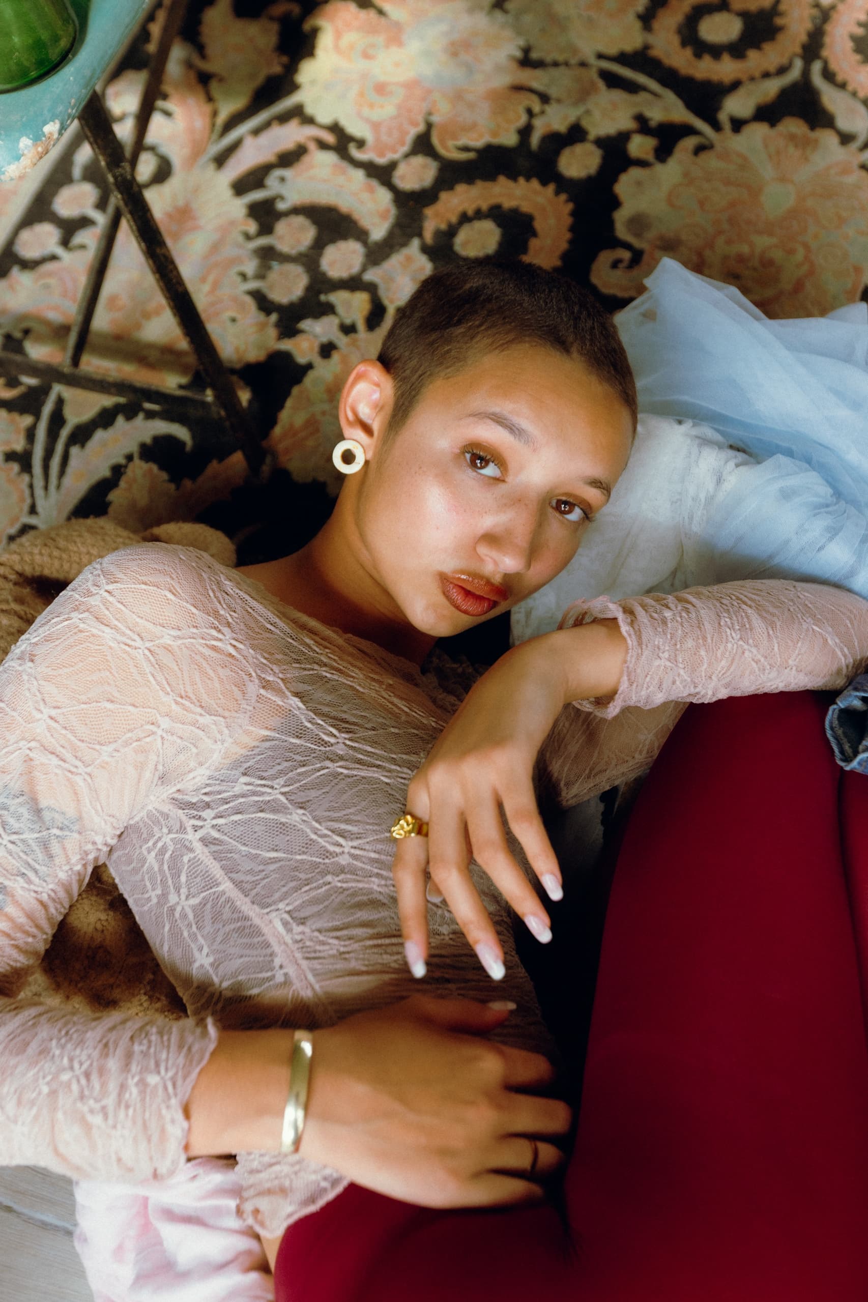 Person lounging on a patterned rug, wearing a lacy top and red pants, with accessories.