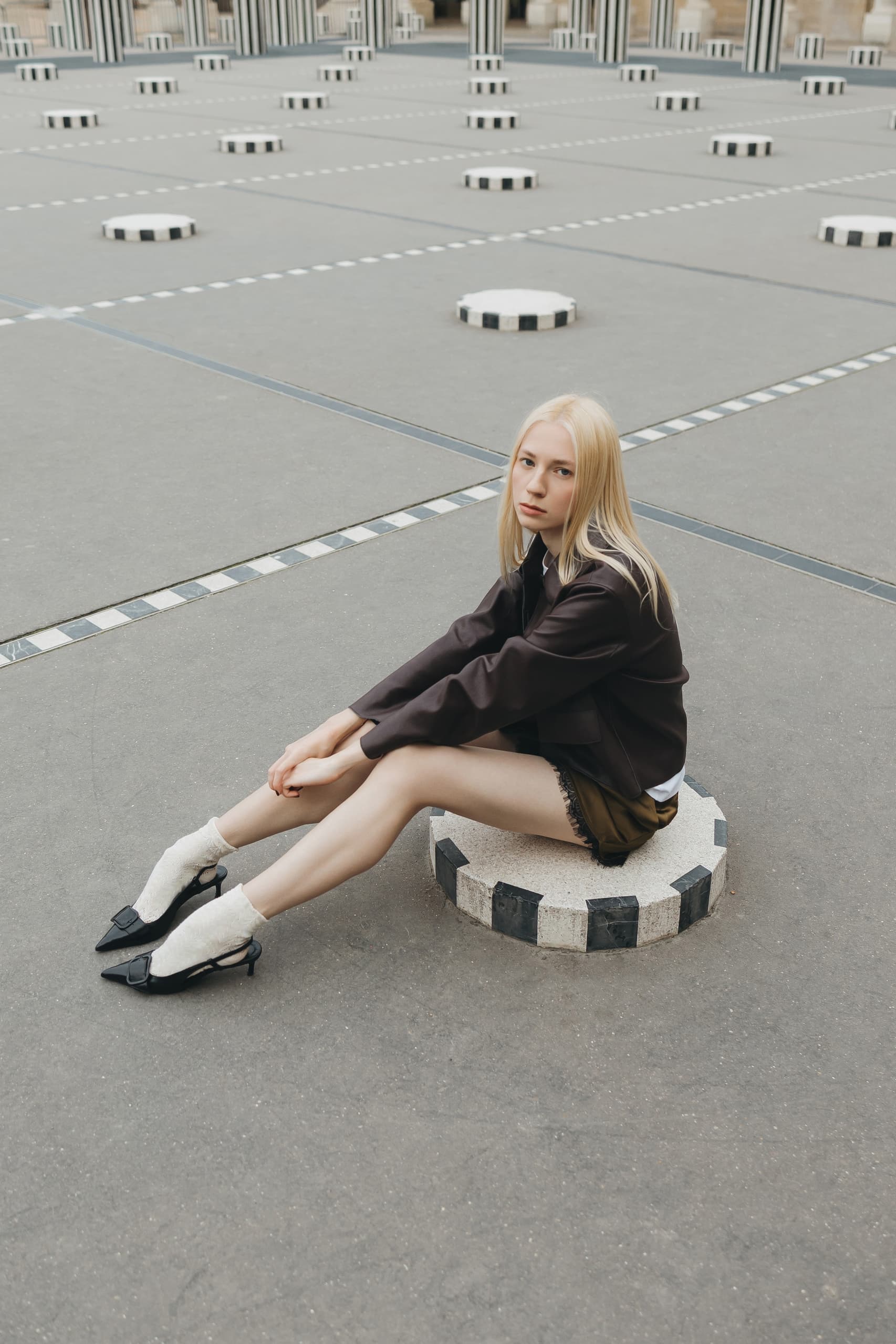 Woman sitting on striped pedestal in courtyard, wearing black jacket and heels.