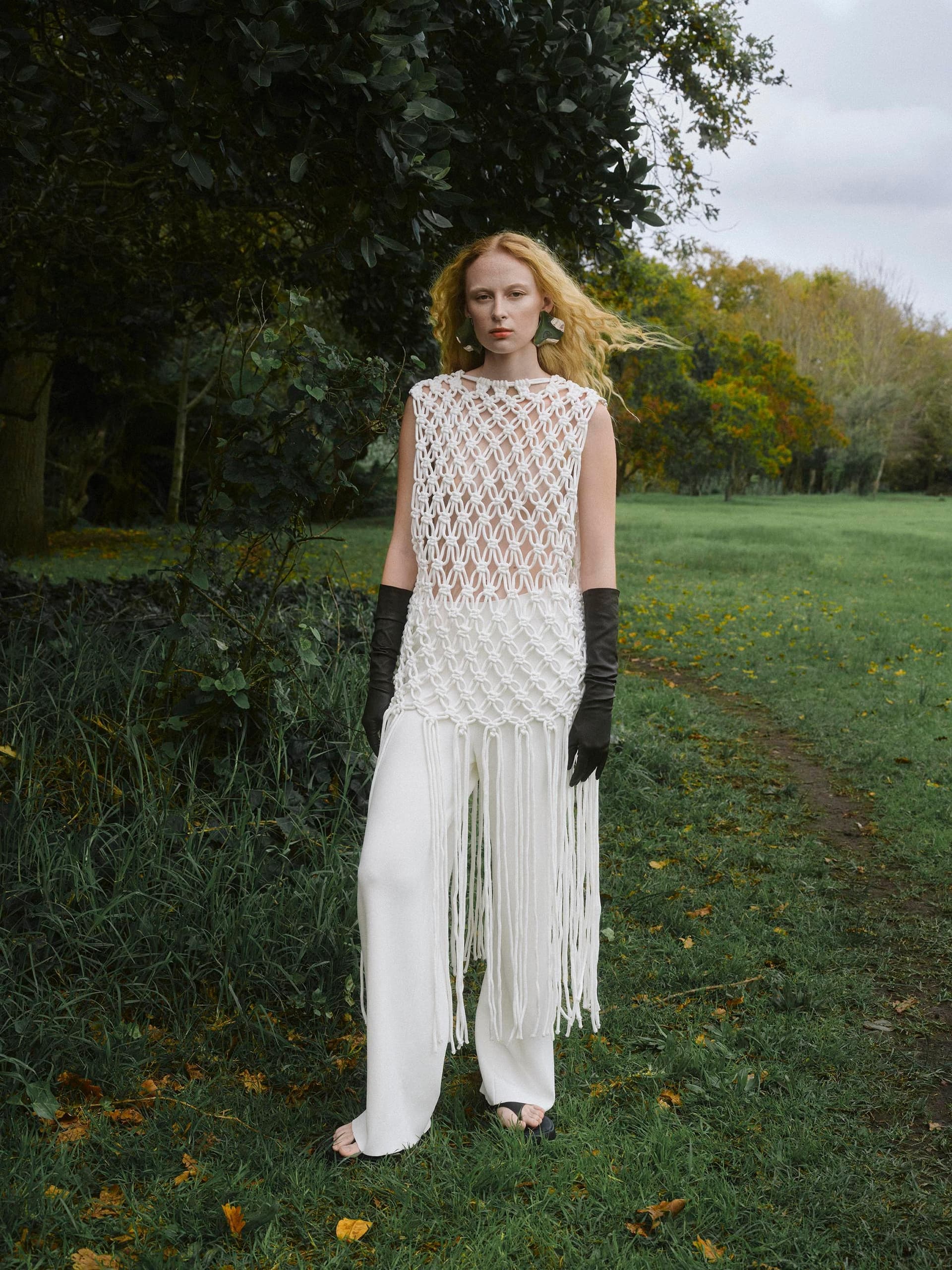 Fashionable woman in white fringe outfit standing in a lush green garden.