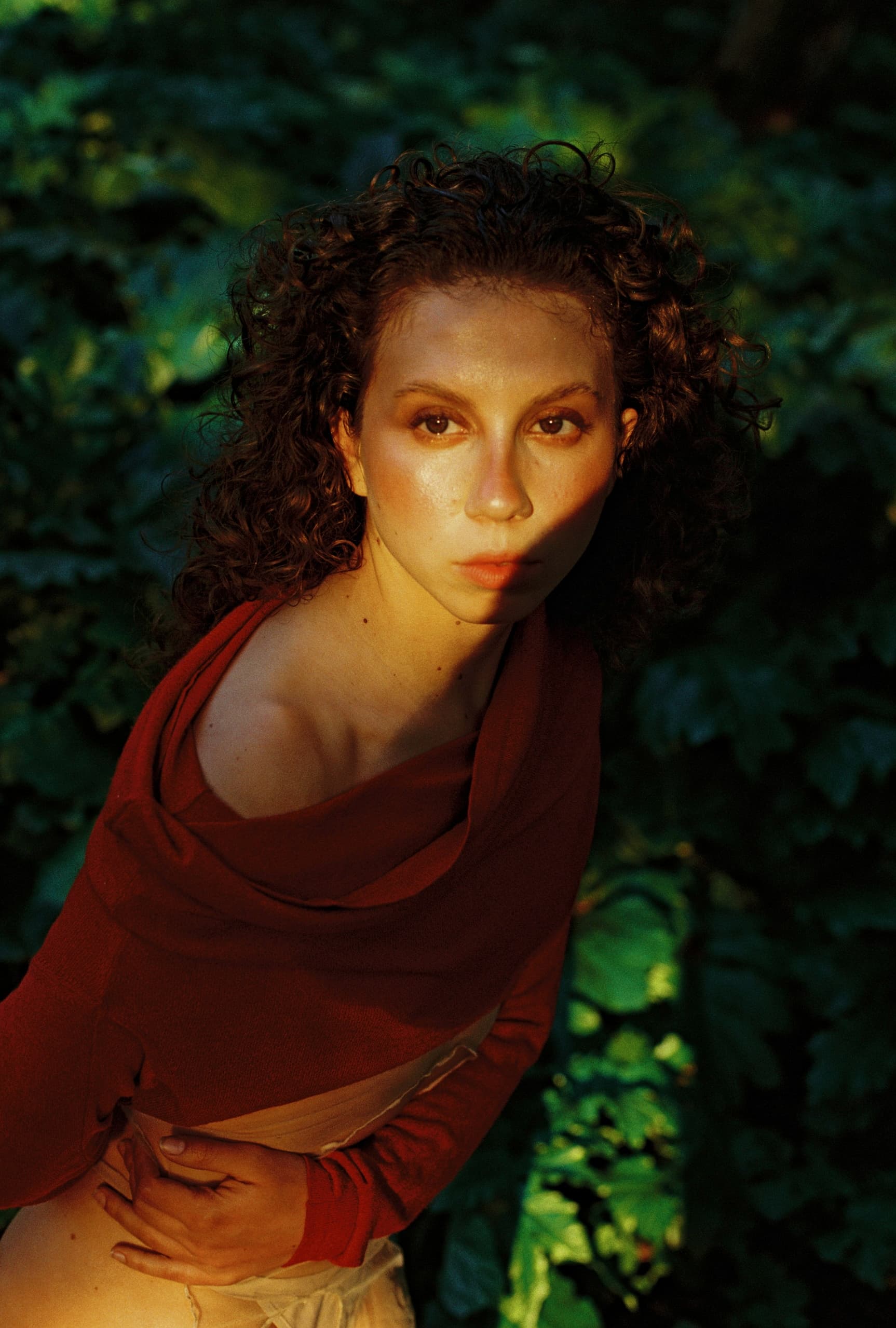 Woman in red shirt stands in sunlit forest, with sunlight on her face.