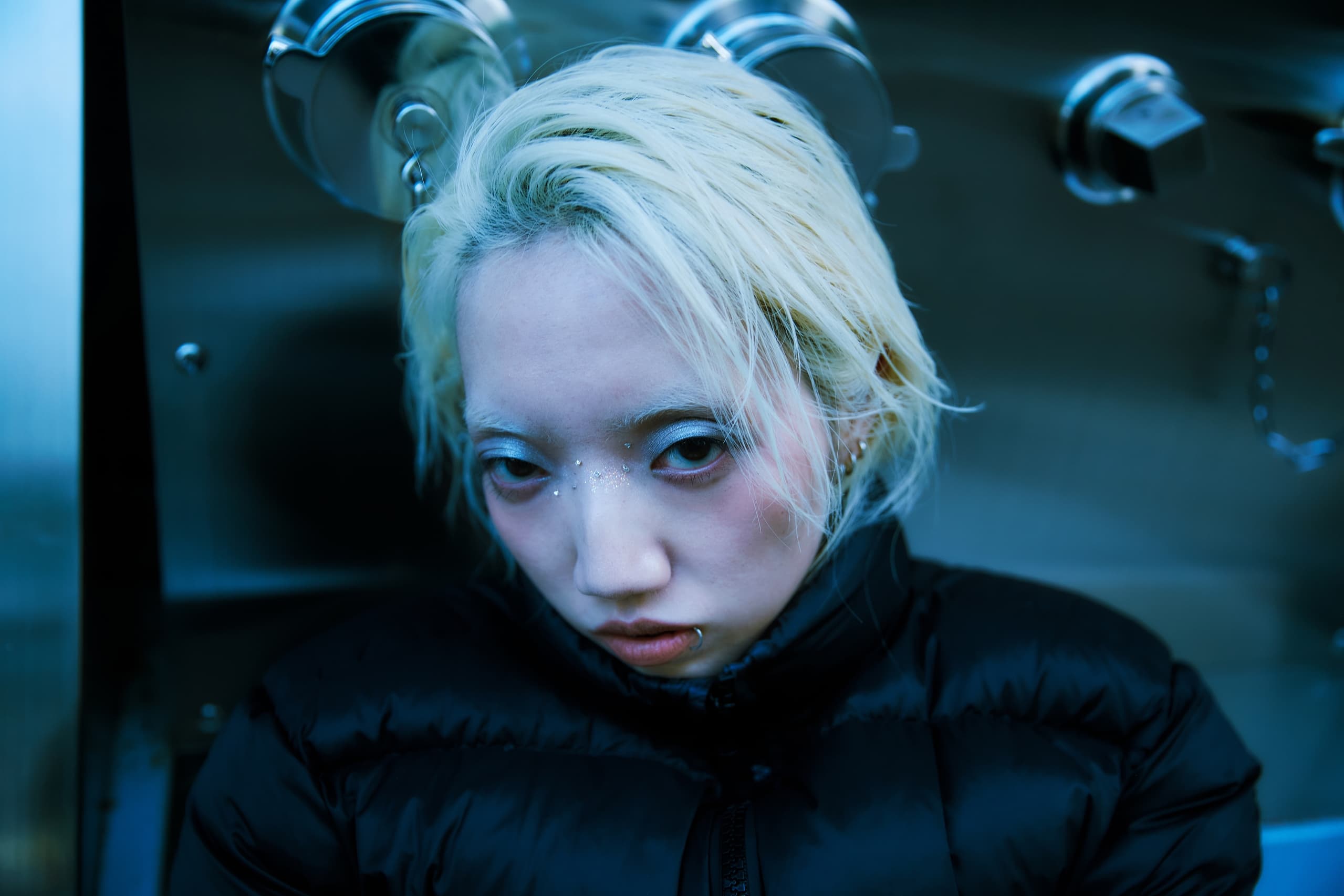 Blonde person in black jacket in industrial setting, piercing gaze.