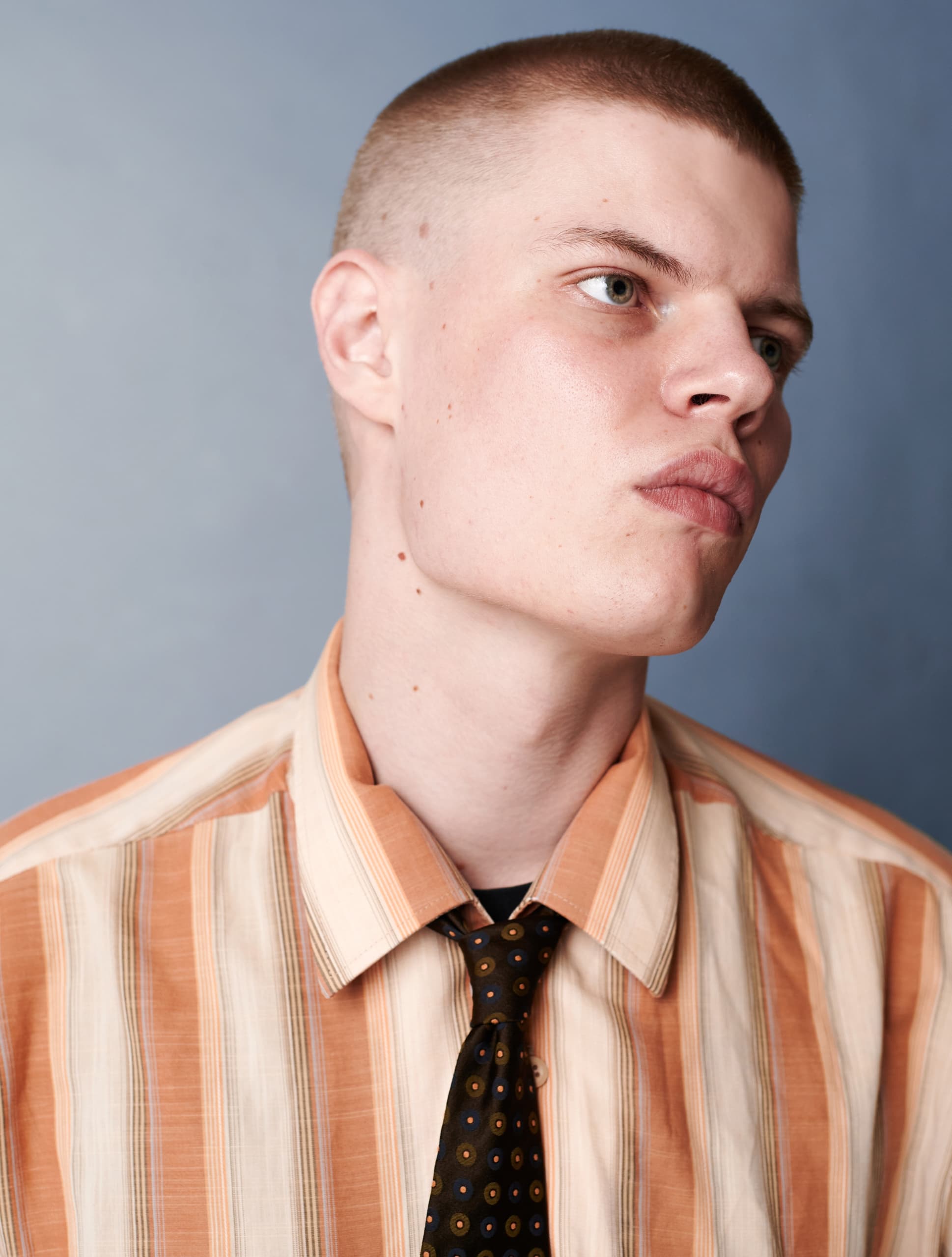 Young man in striped shirt and tie with buzz cut against blue background.