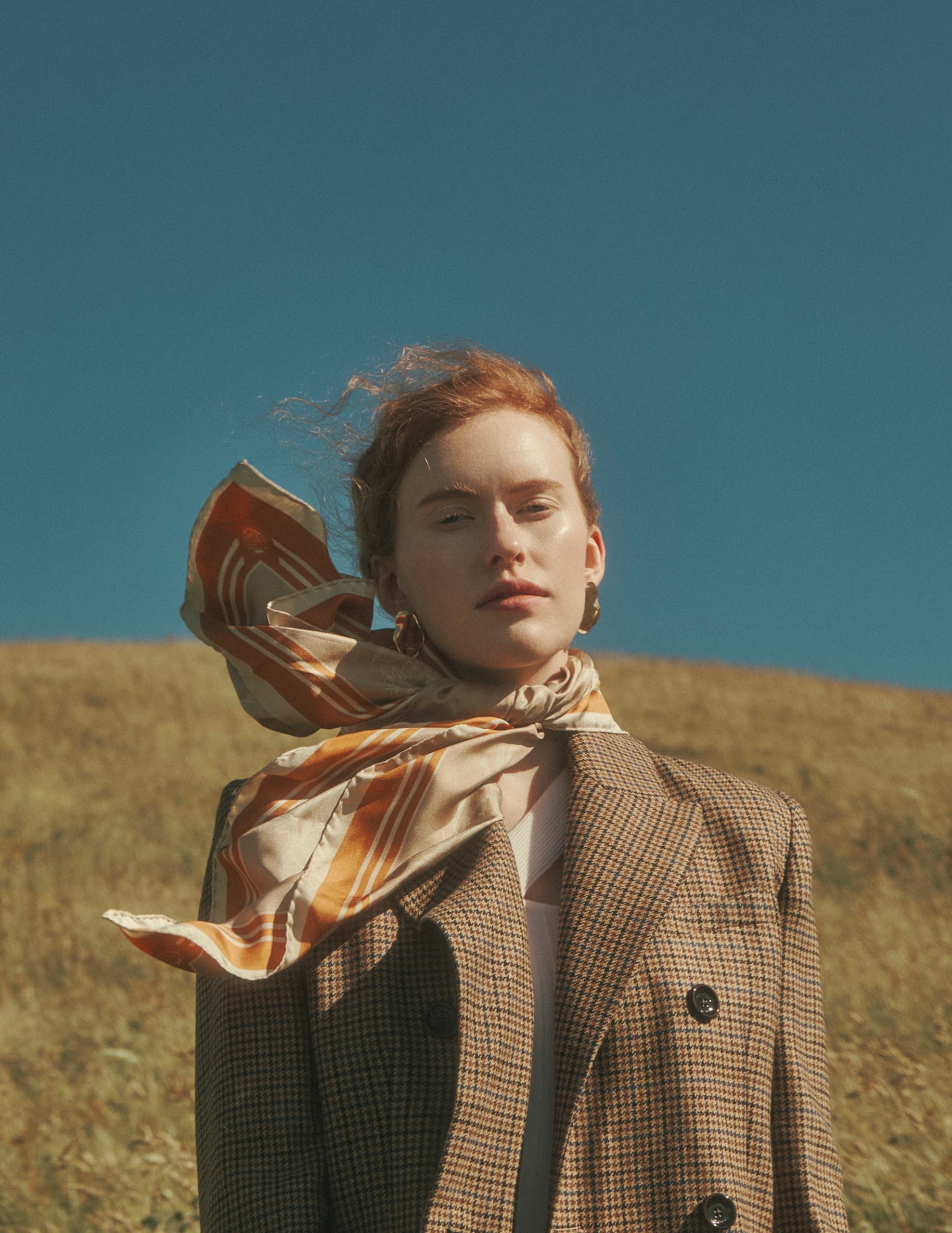 Woman in plaid coat and scarf stands in sunny, grassy field under clear blue sky.