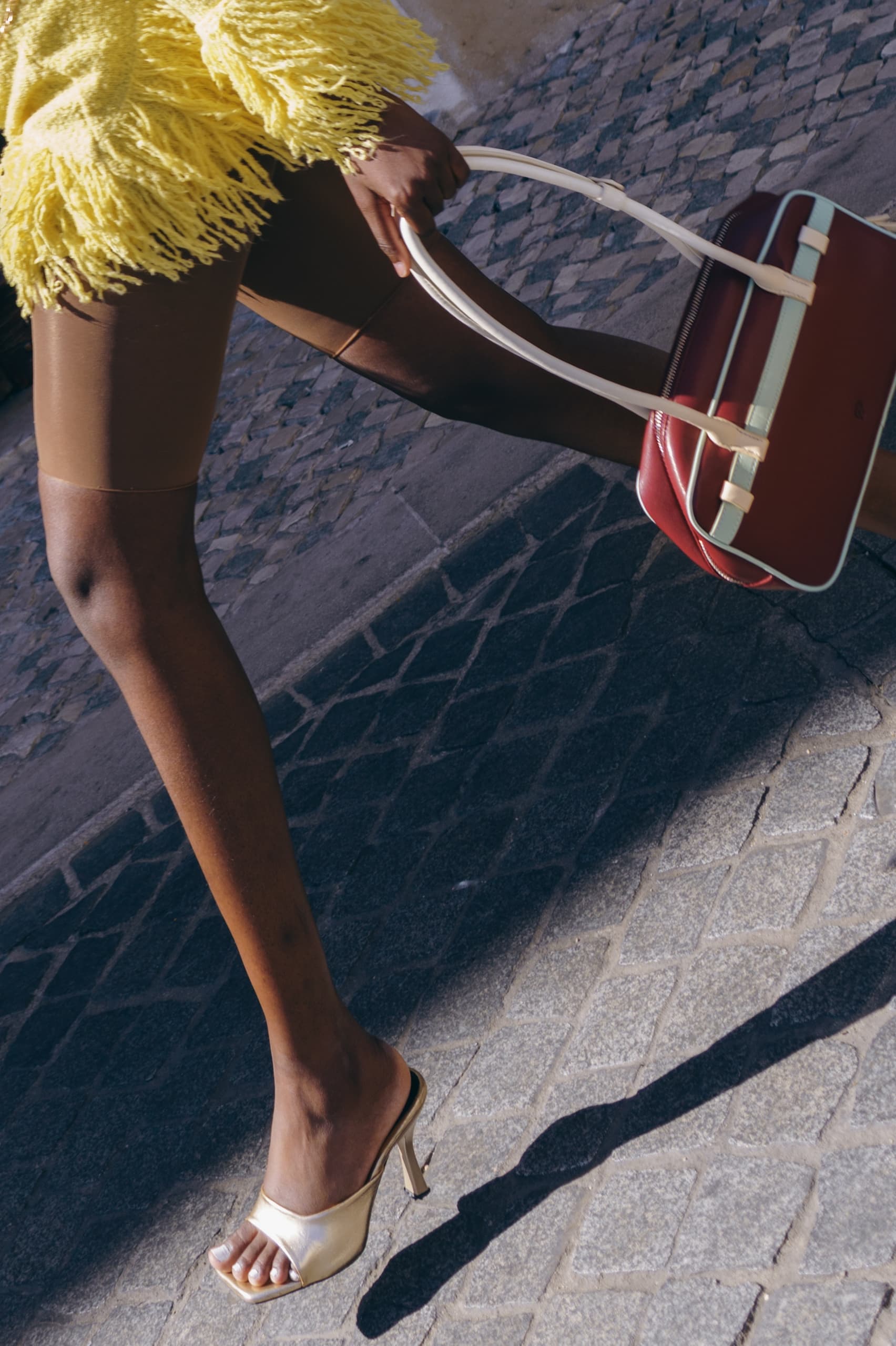 Fashionable stride in heels with yellow fringe dress and stylish handbag on cobblestone.