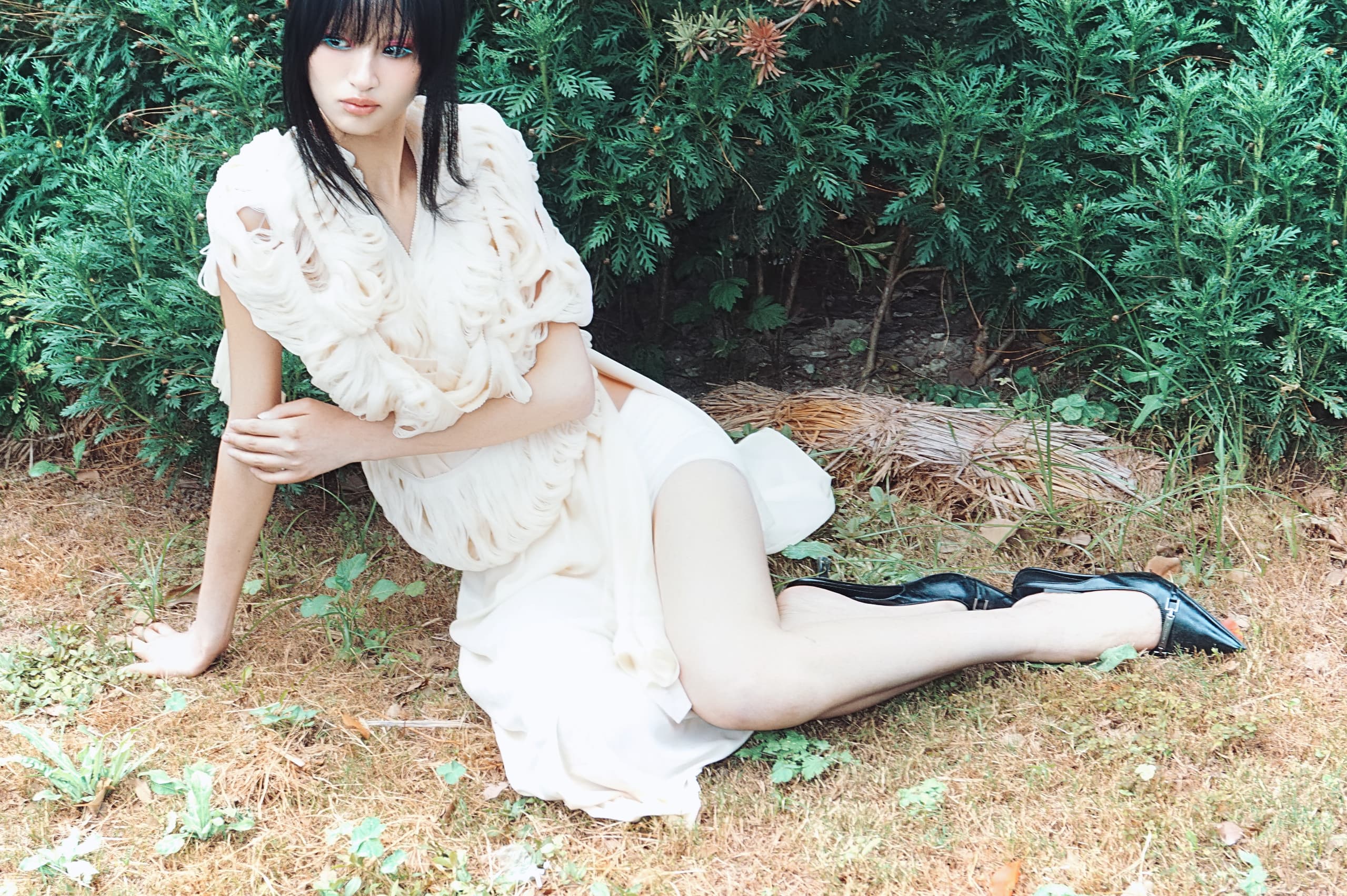 Fashionable woman in white dress sitting on grass beside lush greenery.