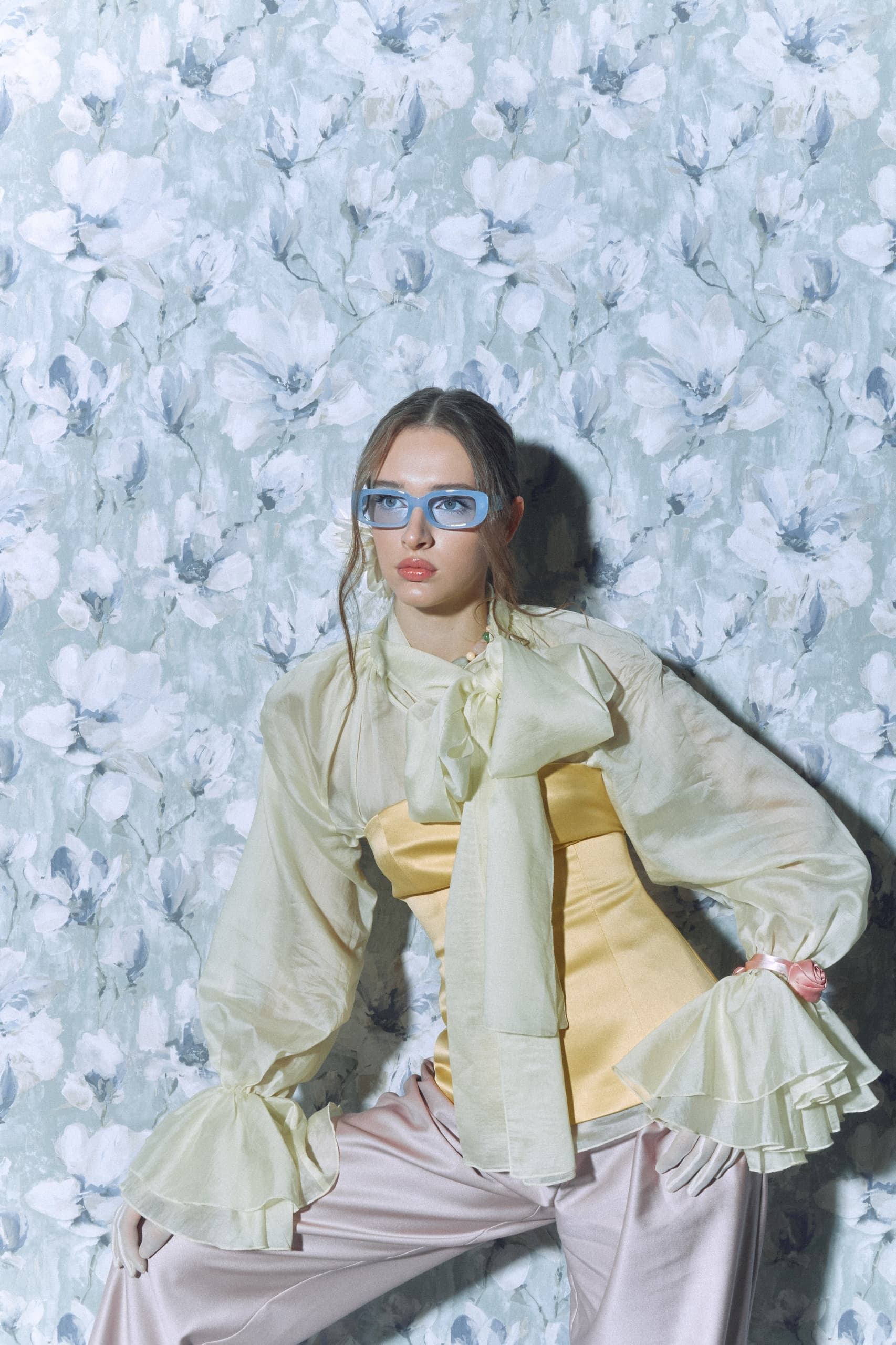 Fashionable woman in pastel outfit with floral background, wearing blue sunglasses.