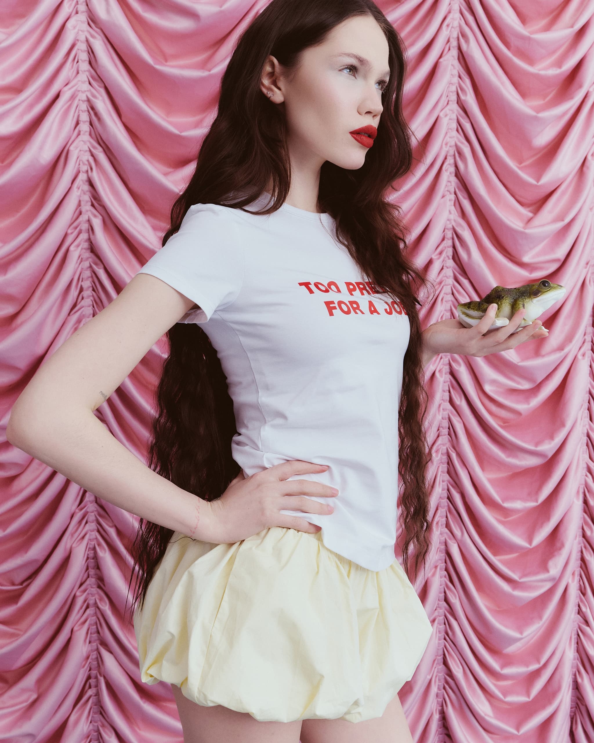 Person with long hair holding a frog, wearing a bold shirt, pink draped backdrop.