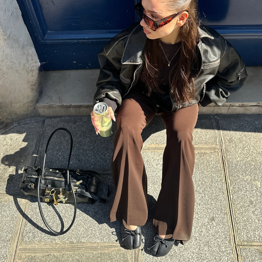 Woman sitting on a step with a drink, stylish outfit and leather bag nearby.