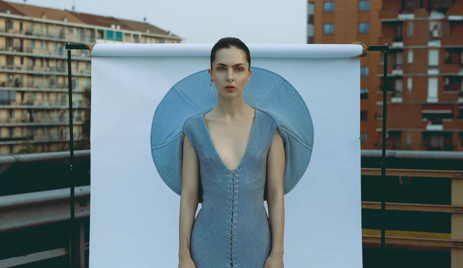 Fashion model in a blue dress with circular design on an urban rooftop backdrop.