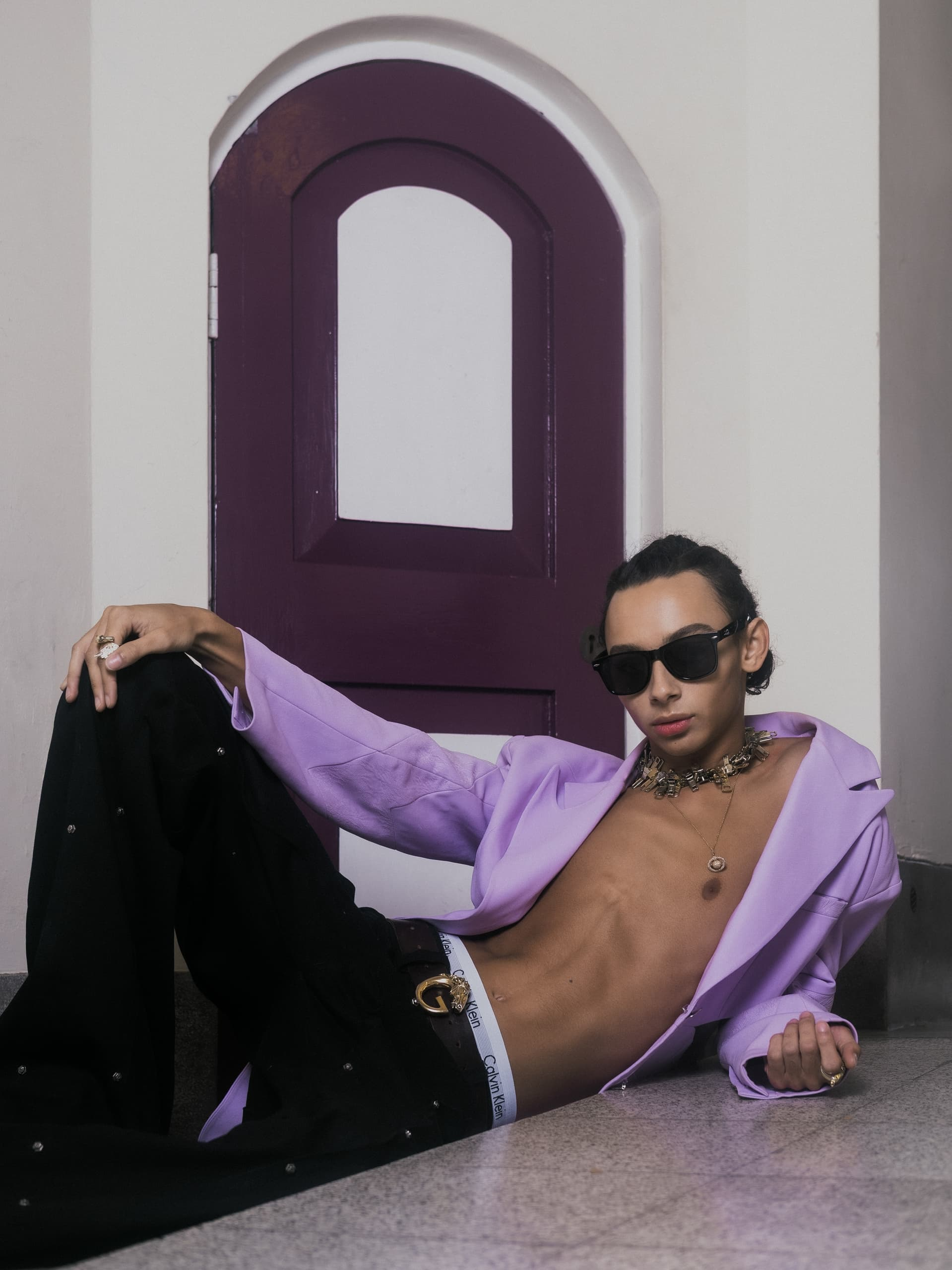 Fashionable person in a lavender jacket, posing stylishly indoors with sunglasses.