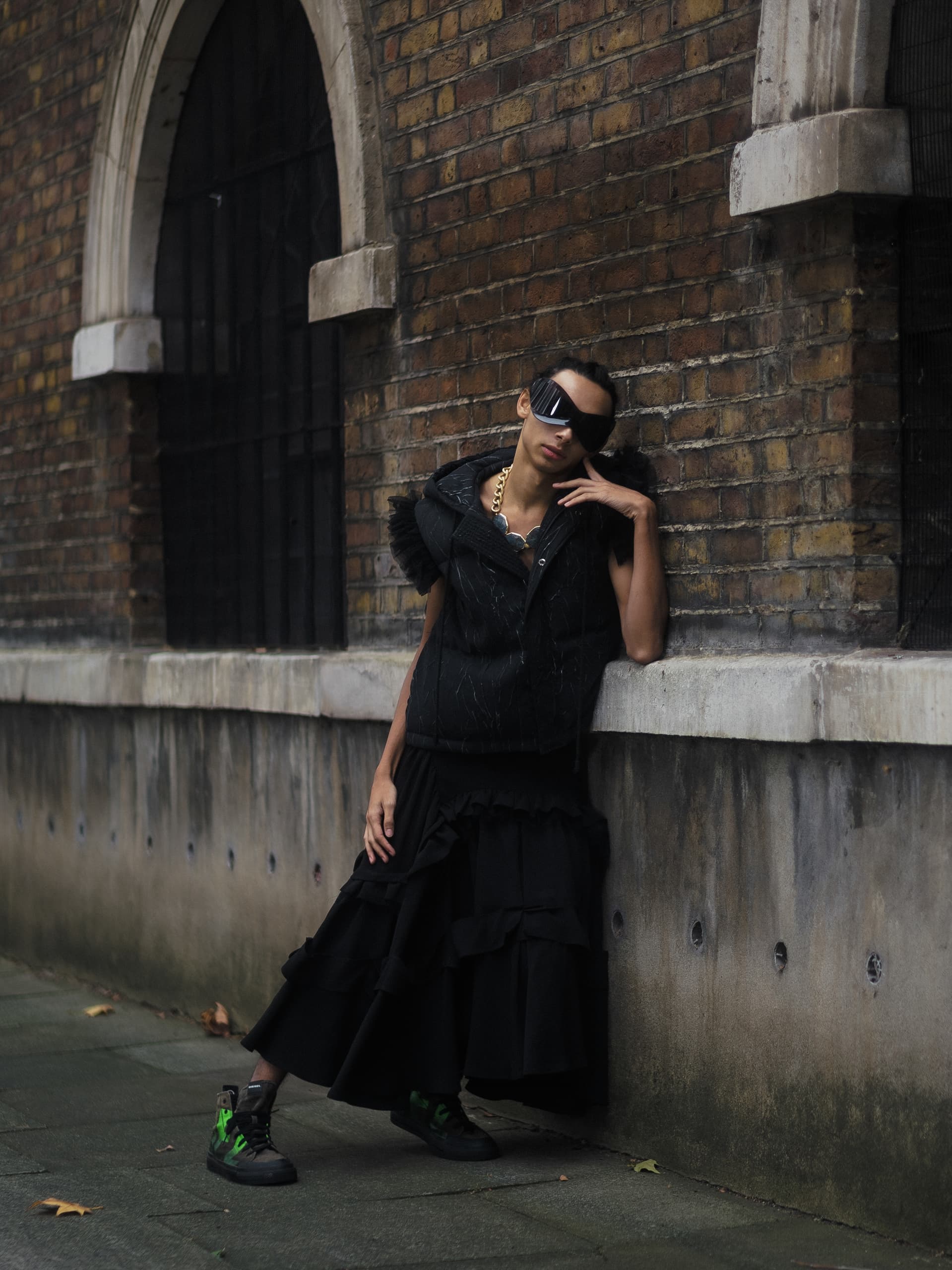 Fashionable person in black outfit and sunglasses leaning against a brick wall.