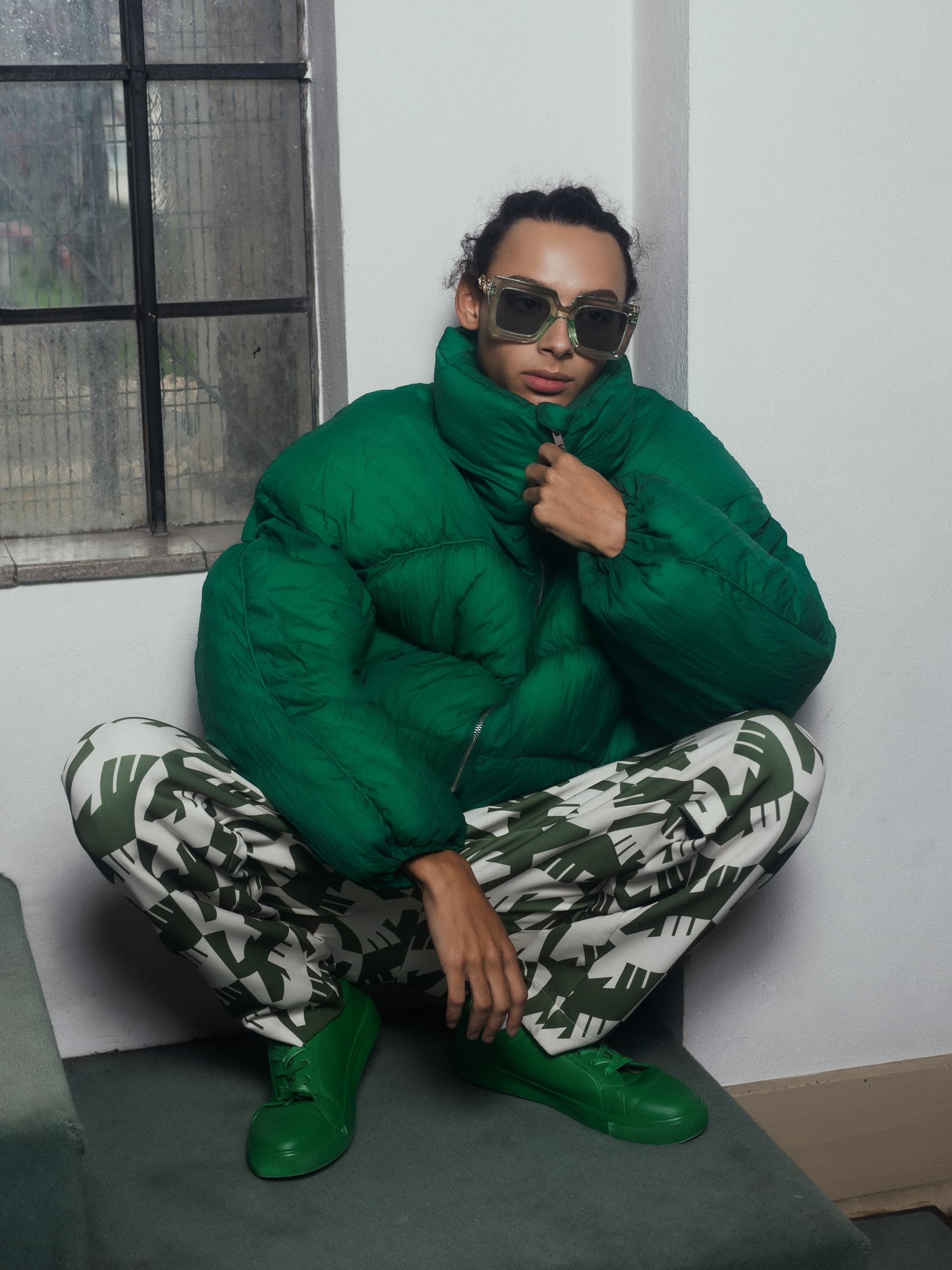 Person in stylish green outfit and sunglasses by window, striking a fashion-forward pose.