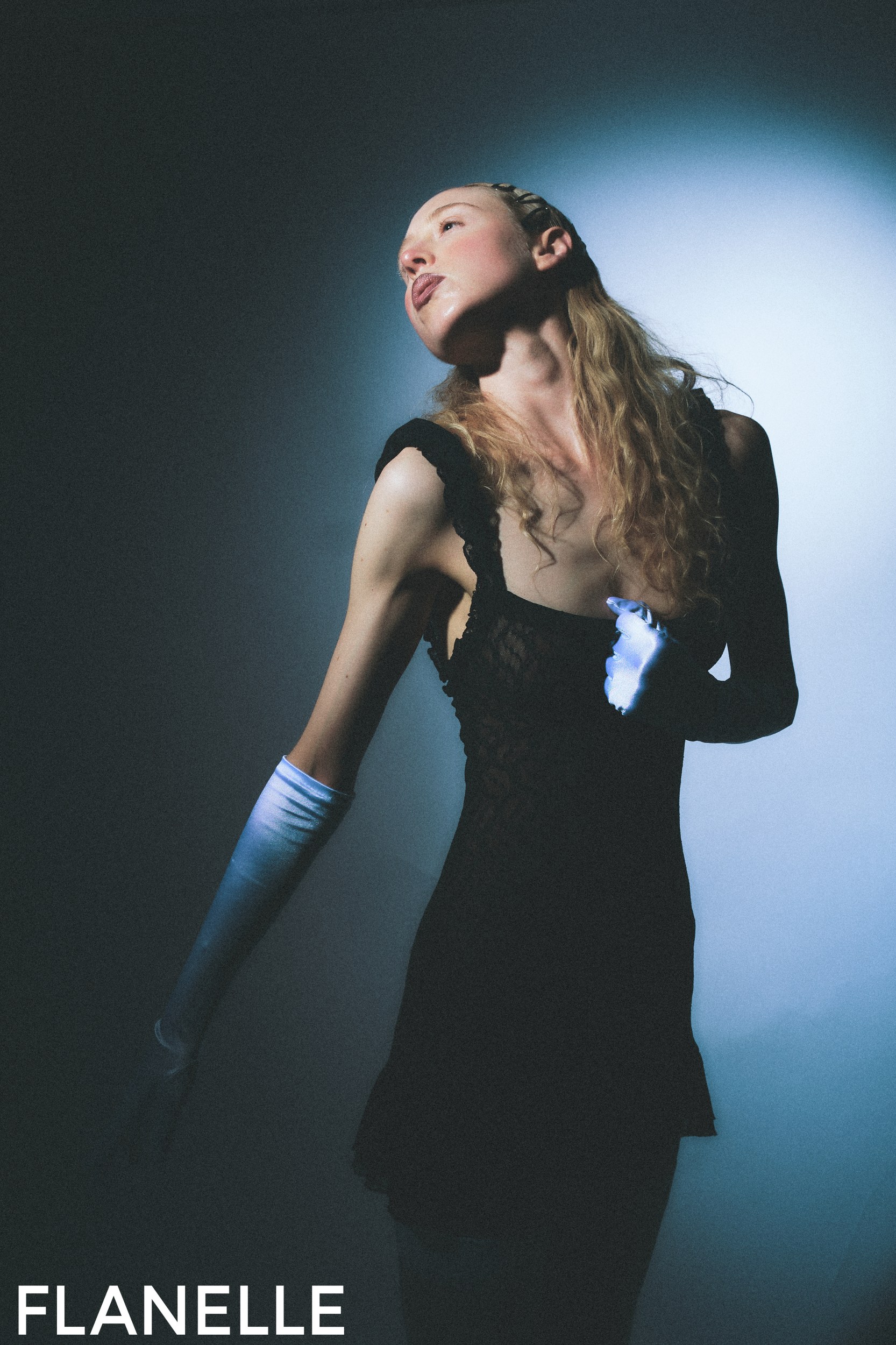 Elegant model in black dress and gloves posing dramatically against a soft spotlight.