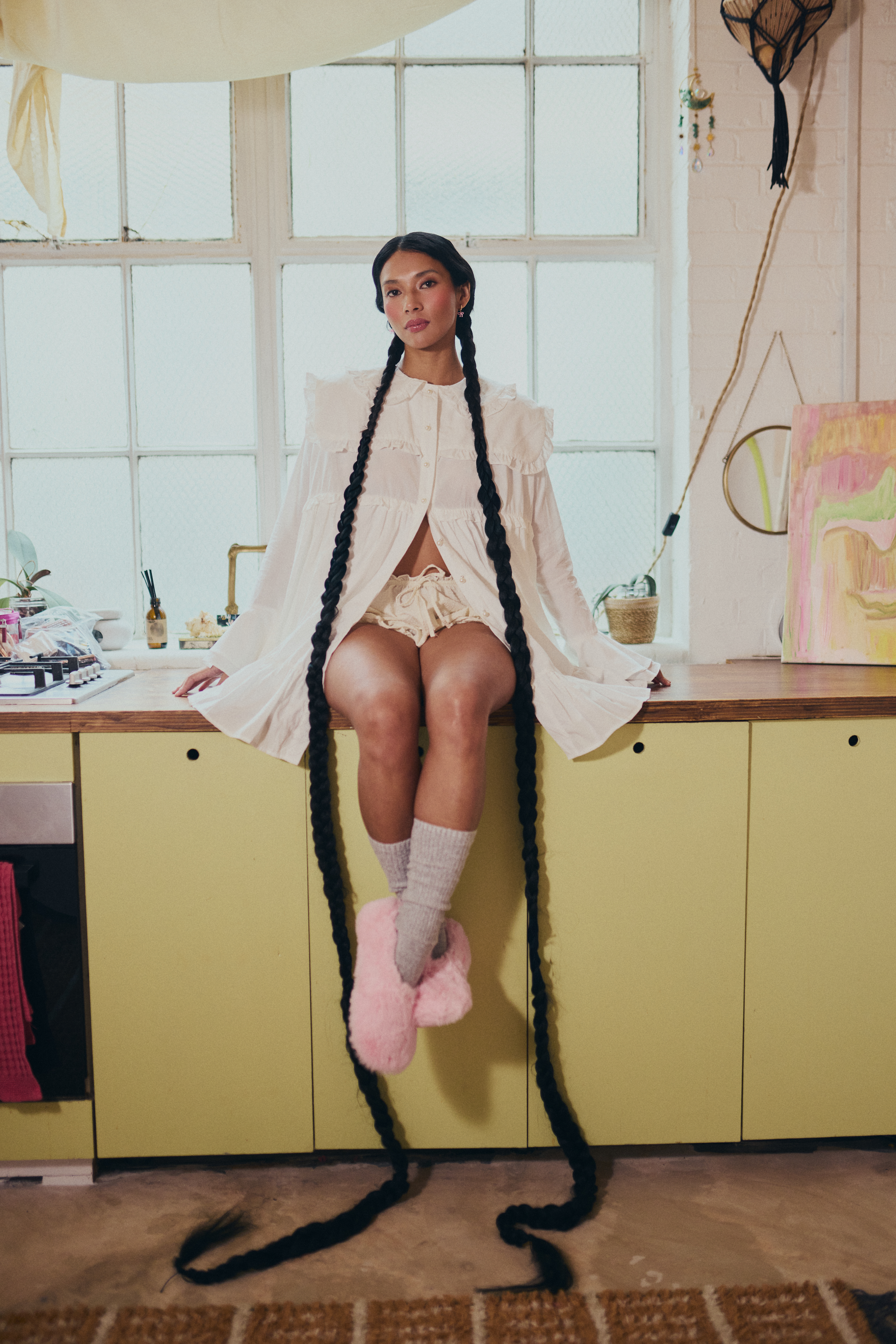 Woman in long braids sitting on kitchen counter, wearing cozy attire and pink slippers.