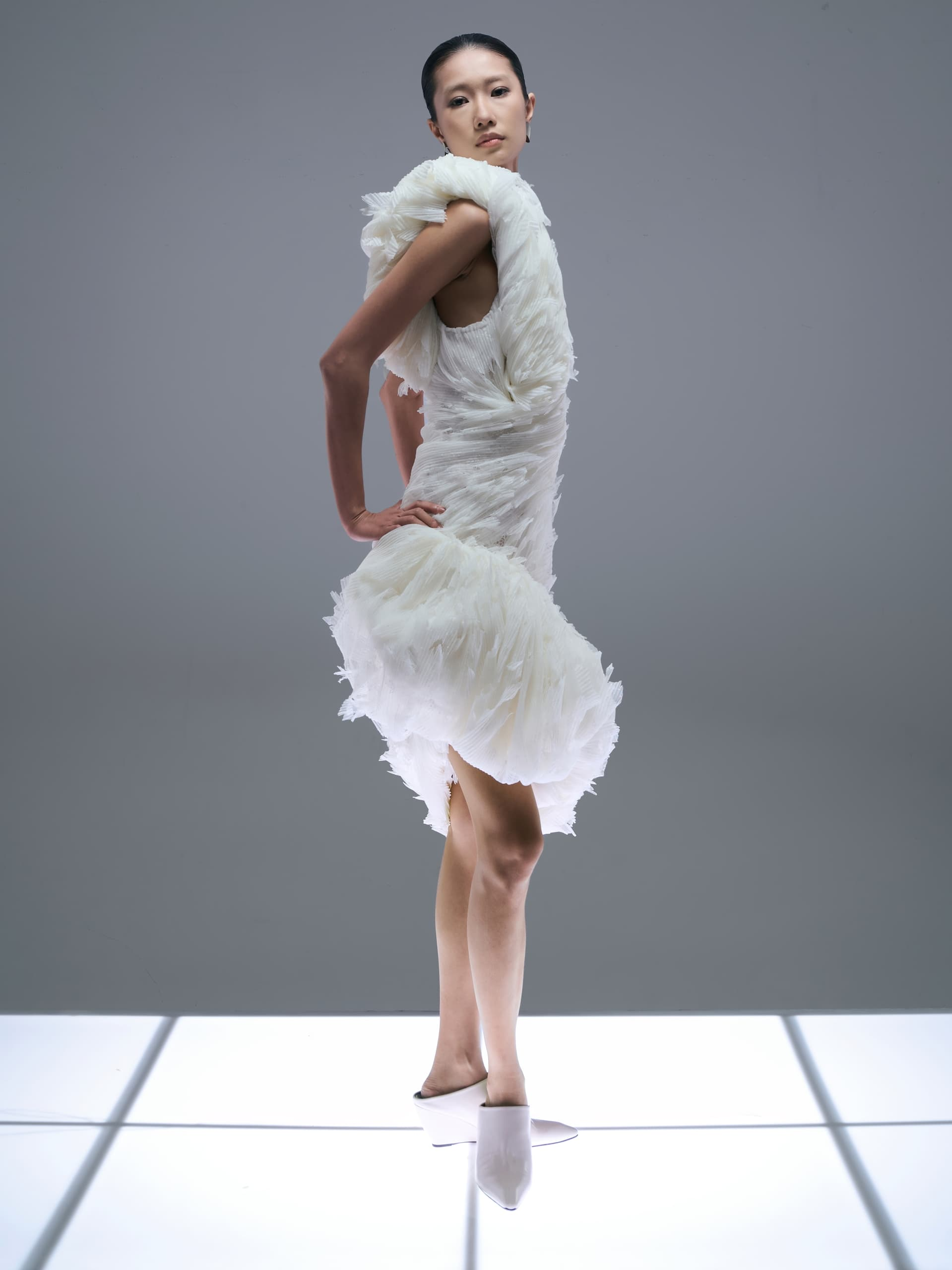 Model in elegant white dress standing on lit floor against gray background.