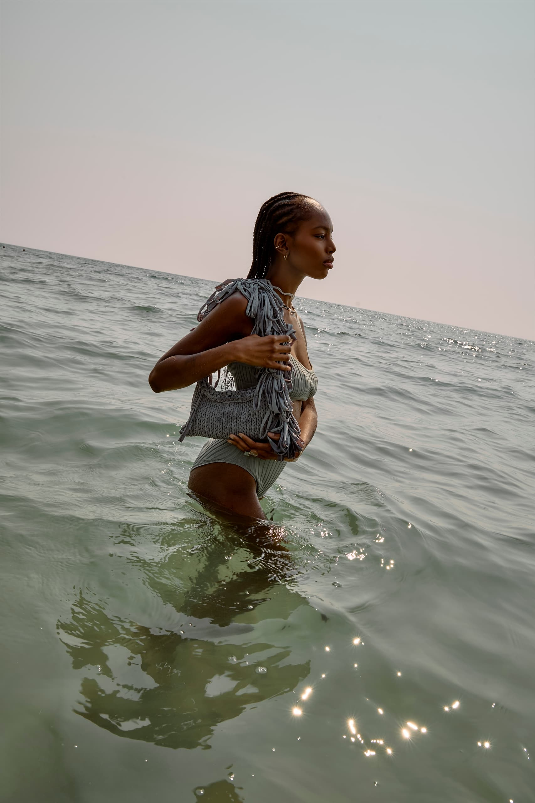 Woman in swimsuit with stylish fringe bag walking in the ocean at sunset.