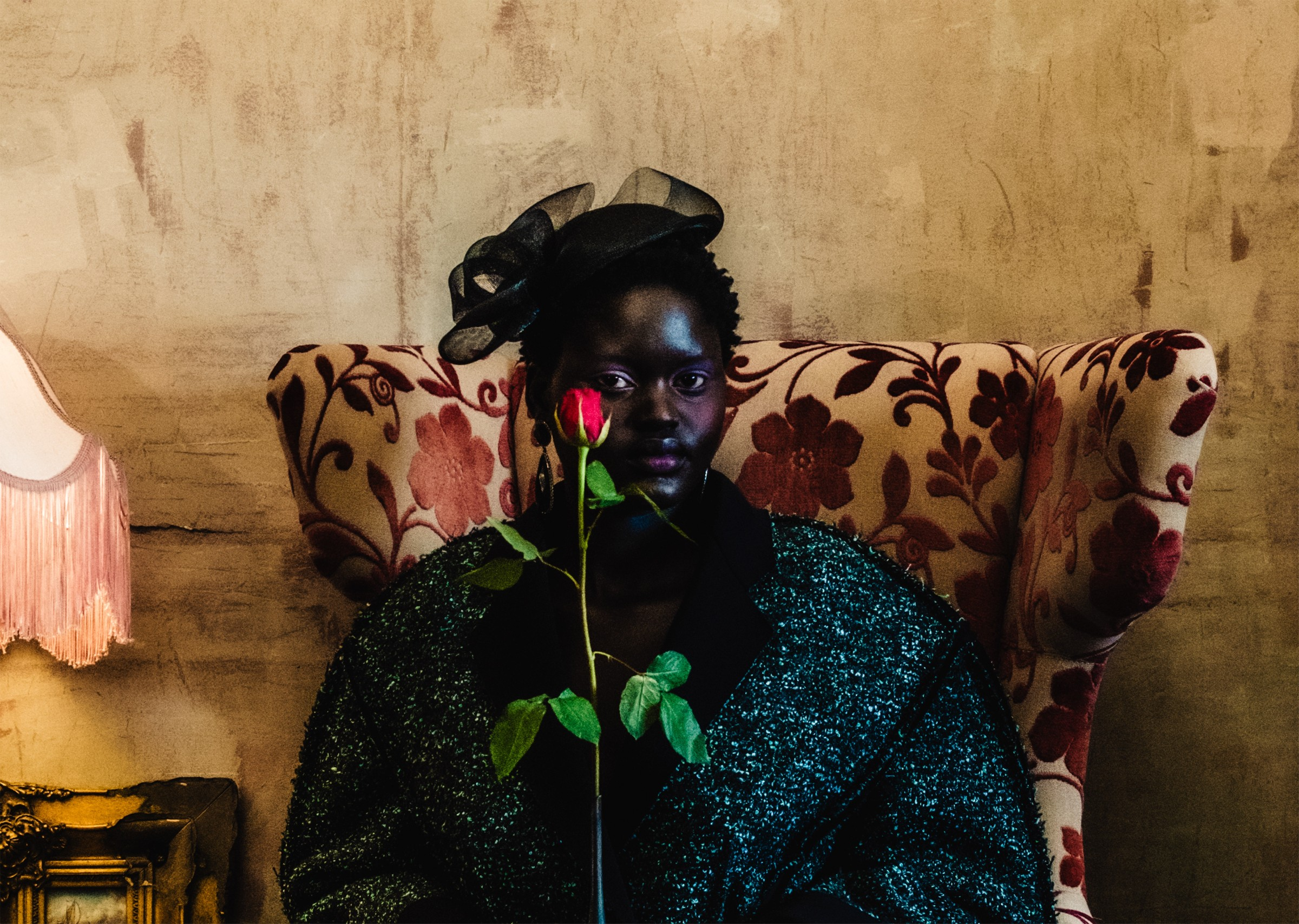 Person holds a red rose, sitting on a floral armchair, vintage setting.