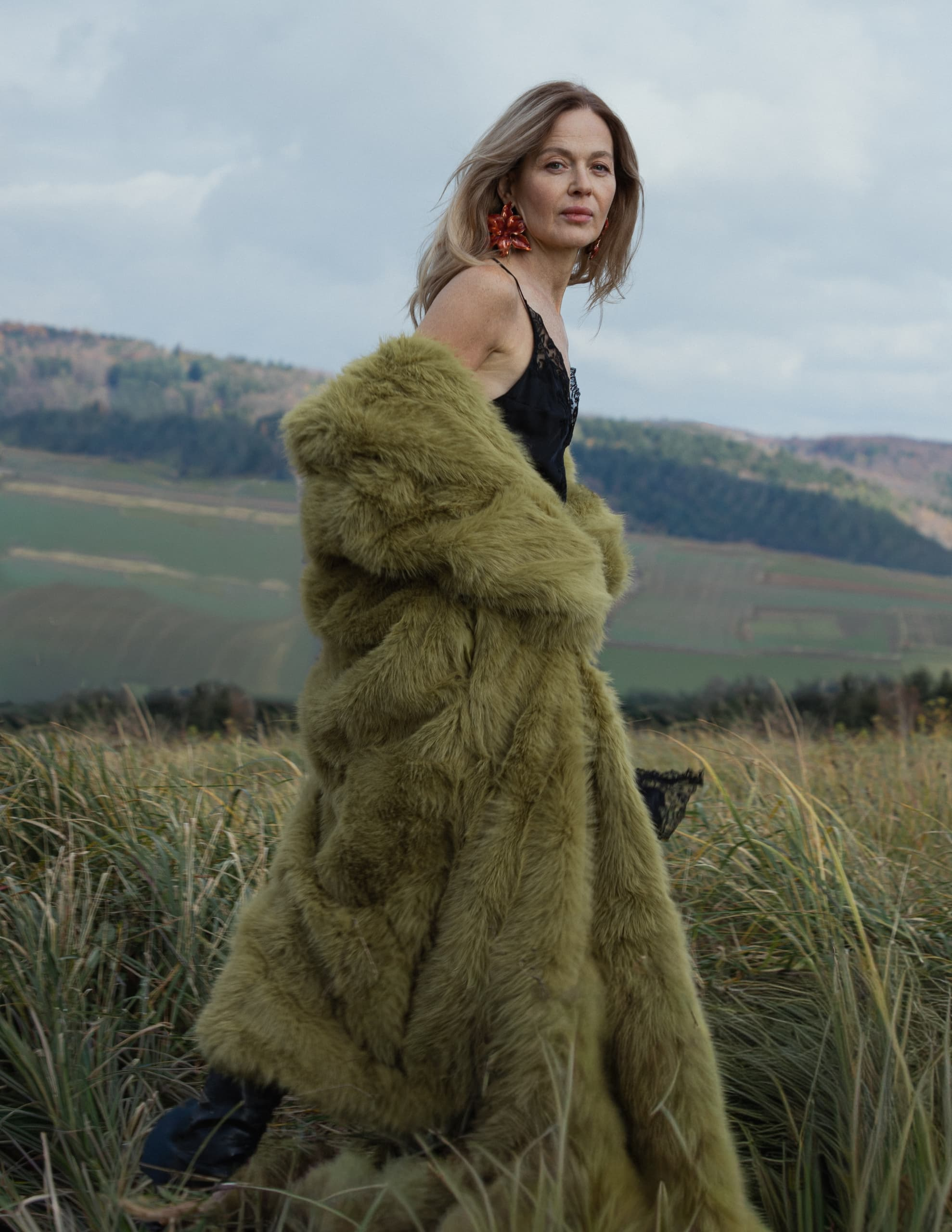 Woman in a green fur coat standing in a scenic field landscape.