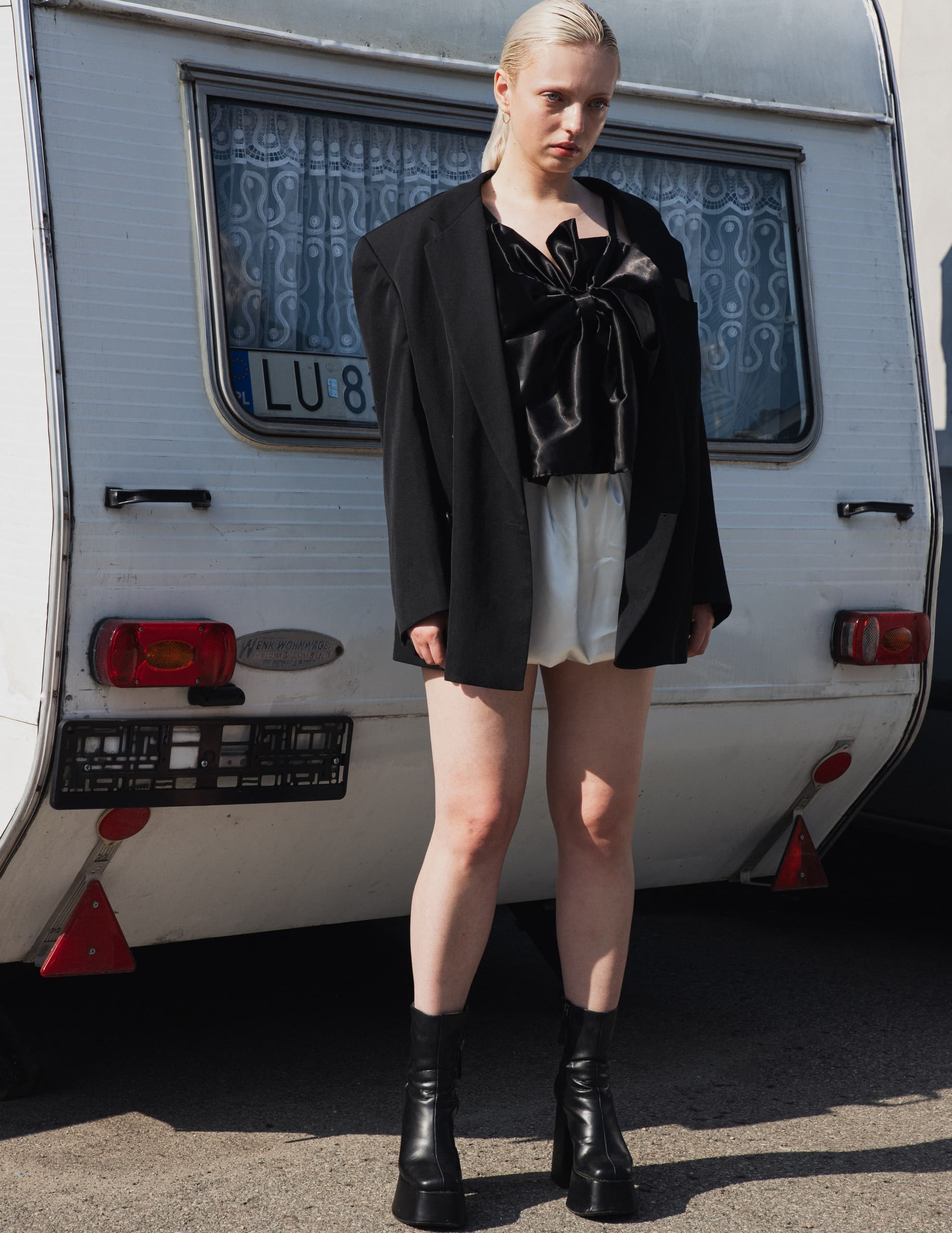 Fashionable person in black blazer and boots standing by a vintage caravan.