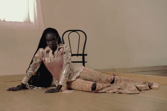 Model in floral fashion on wooden floor with chair backdrop.