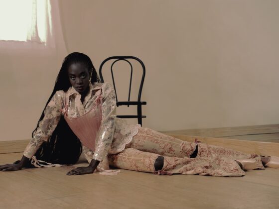 Model in floral fashion on wooden floor with chair backdrop.