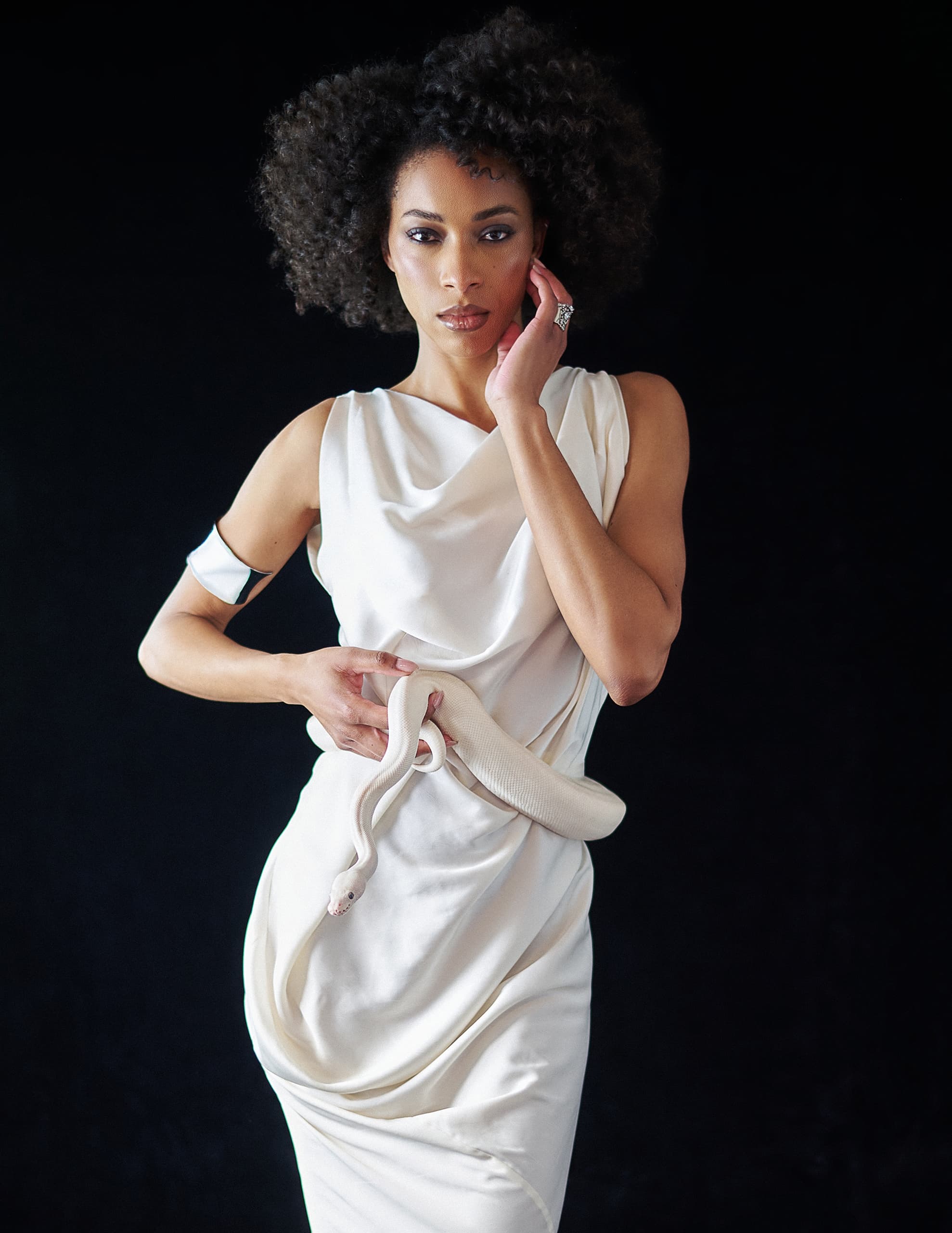 Woman in white dress holds white snake, posing against a black background.