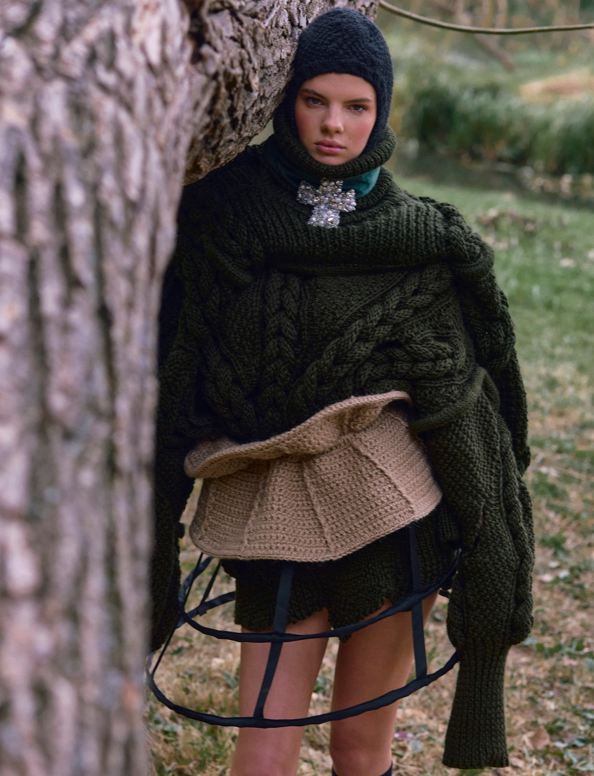 Person in layered knit fashion stands by tree outdoors.