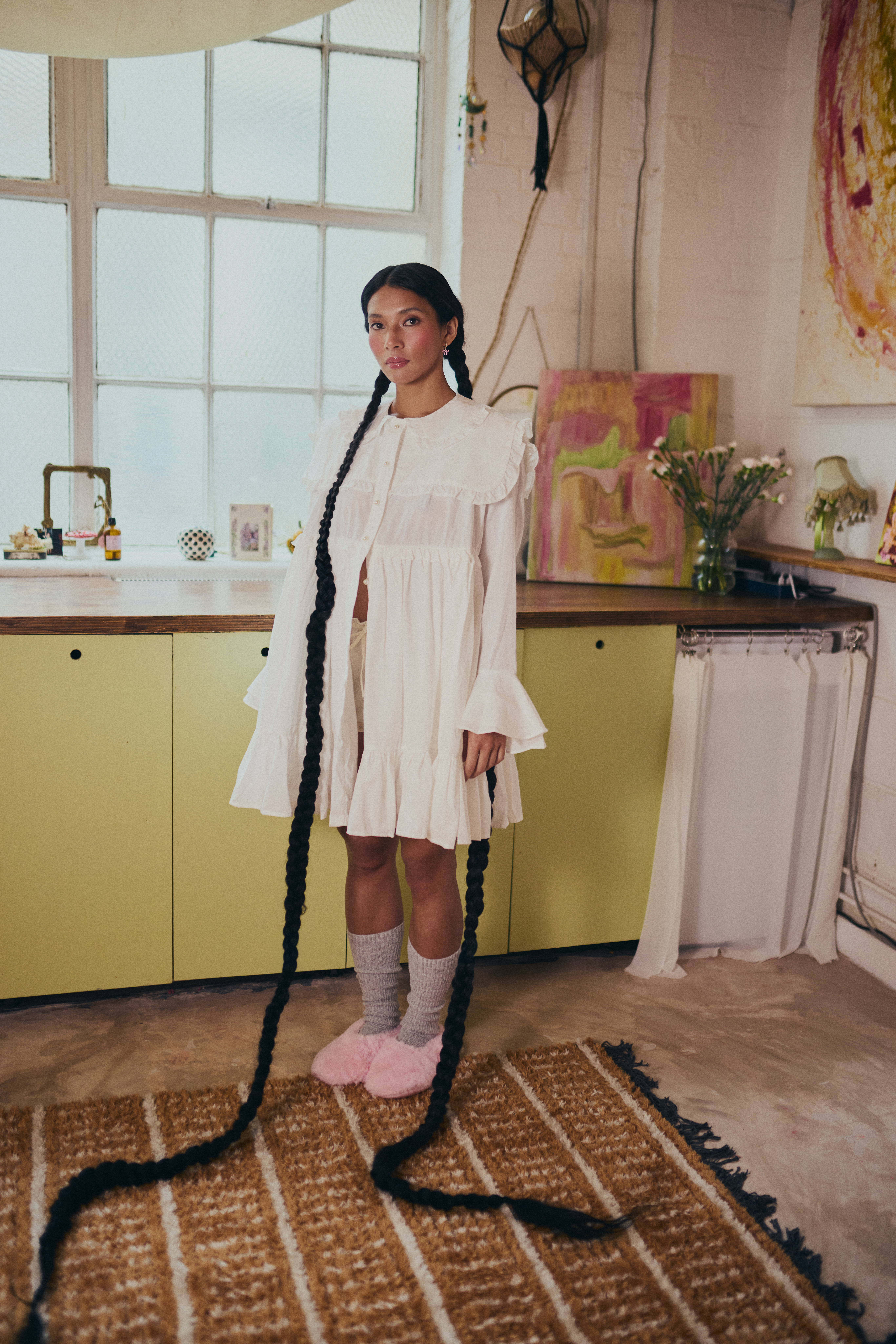 Woman in white dress with long braided hair standing in cozy, art-filled room.