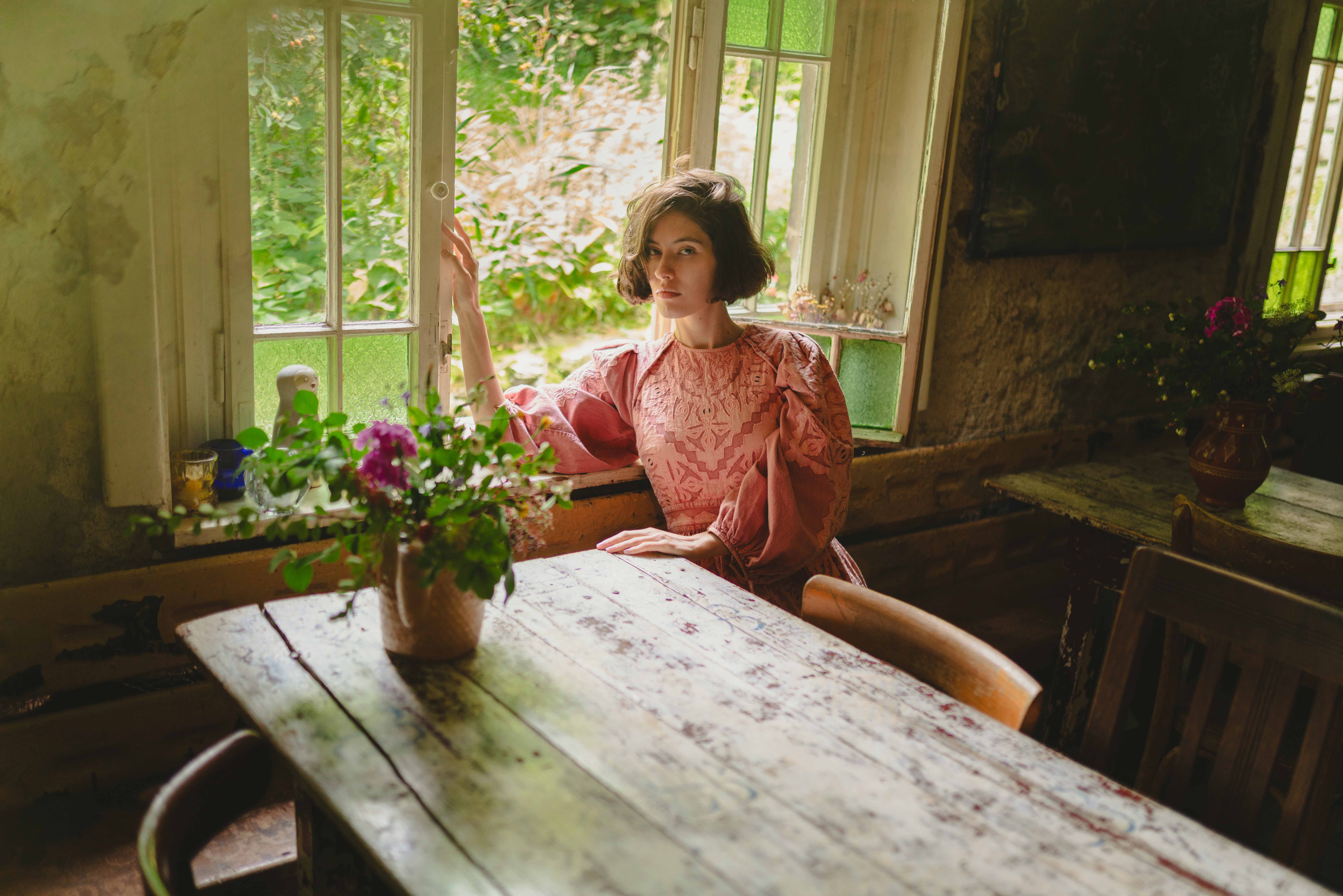 Woman in pink dress gazing out a rustic window with lush greenery outside.