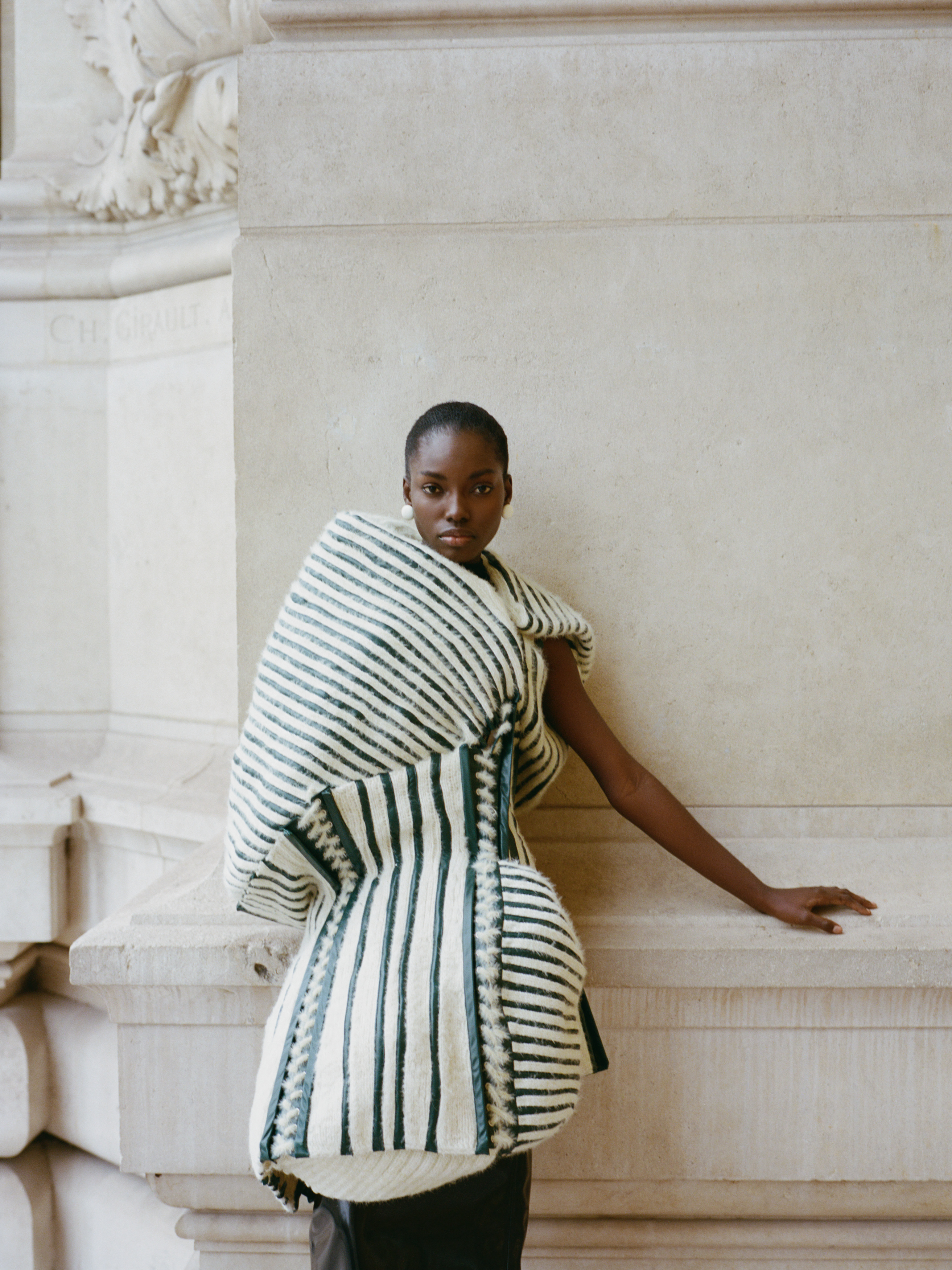 Model wearing a striped, avant-garde outfit, leaning against a stone wall.