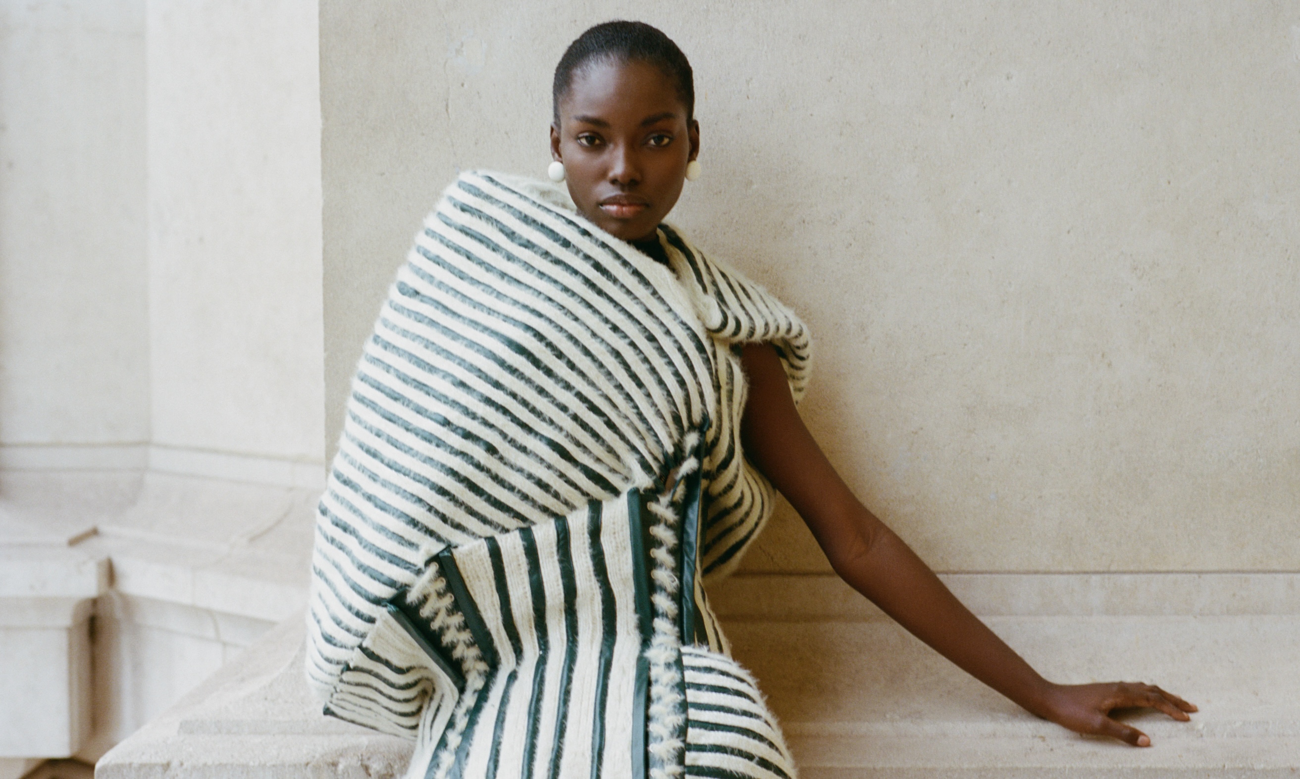 Fashionable person in striped fur garment, leaning on a stone wall.