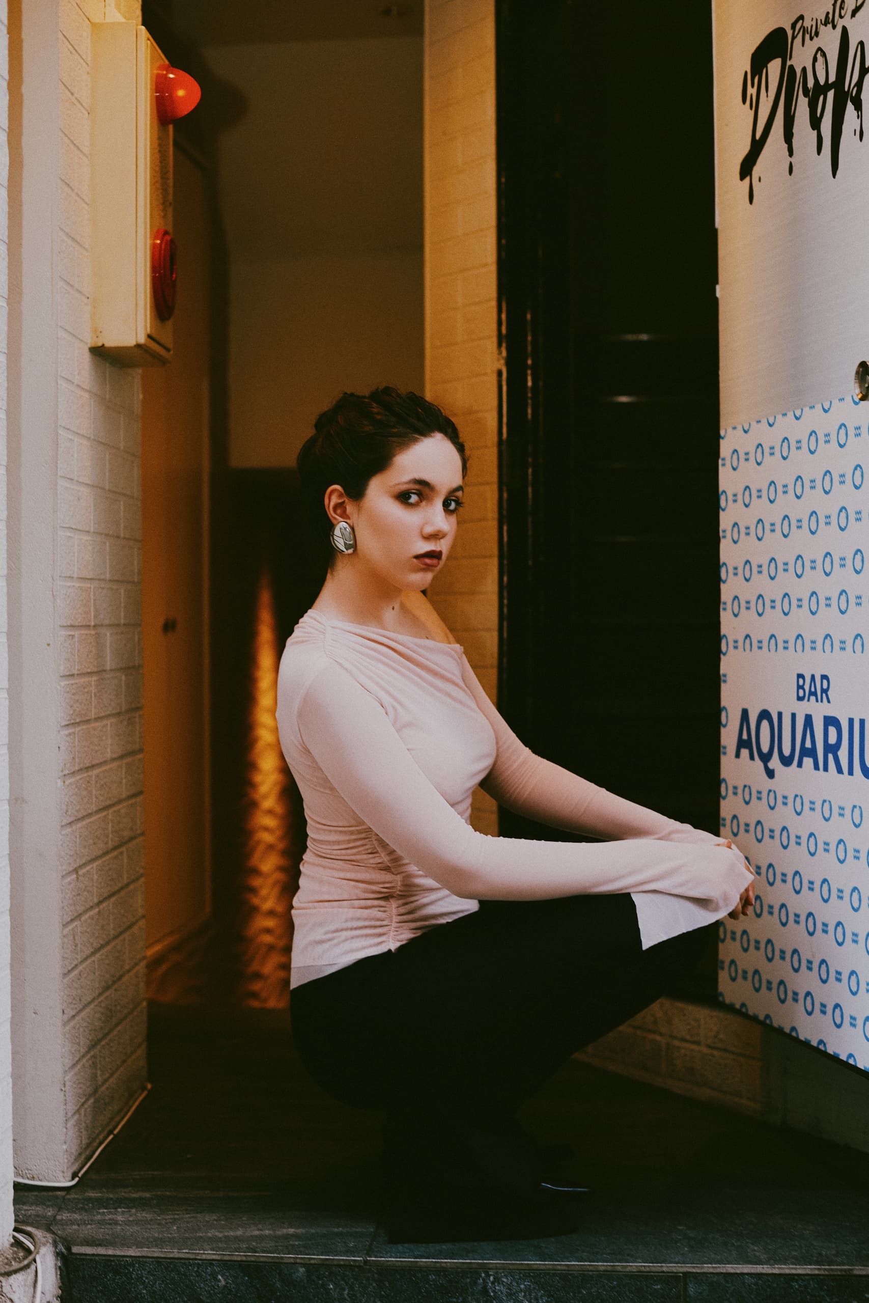 Woman in stylish outfit squats near a sign at a cozy bar entrance.