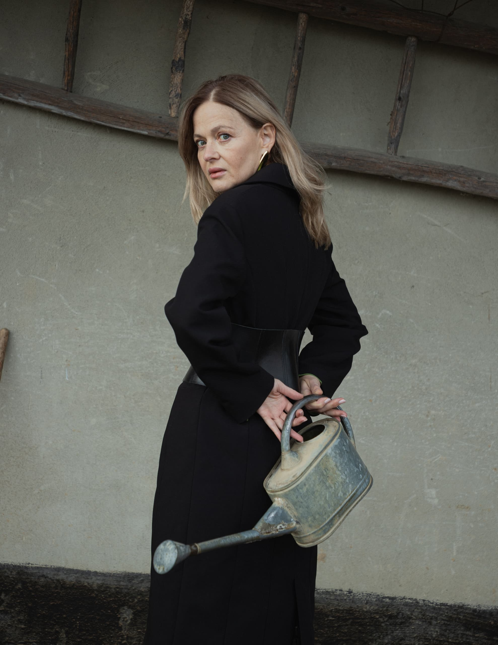 Woman in black coat holding a watering can behind her back, vintage setting.
