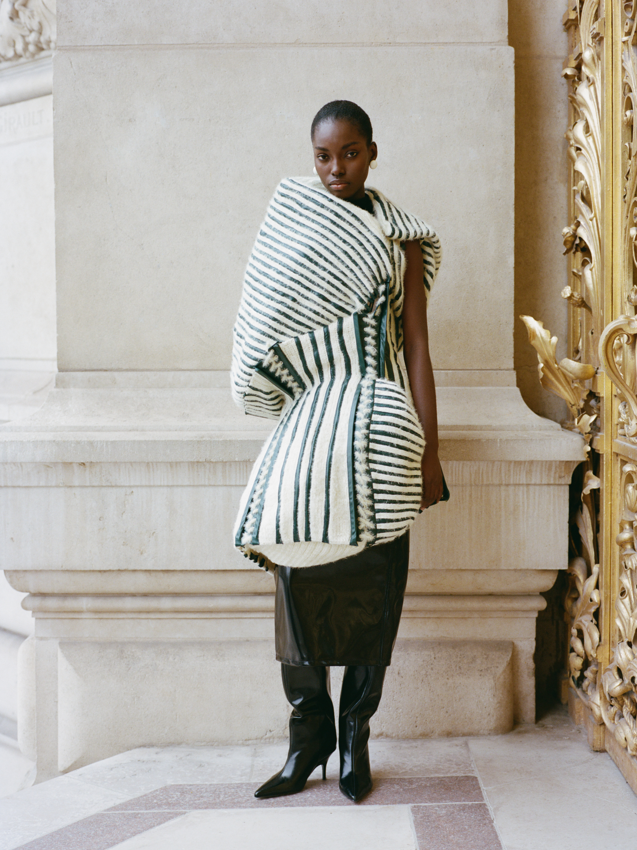 Fashion model in a striped avant-garde coat and boots near ornate architecture.