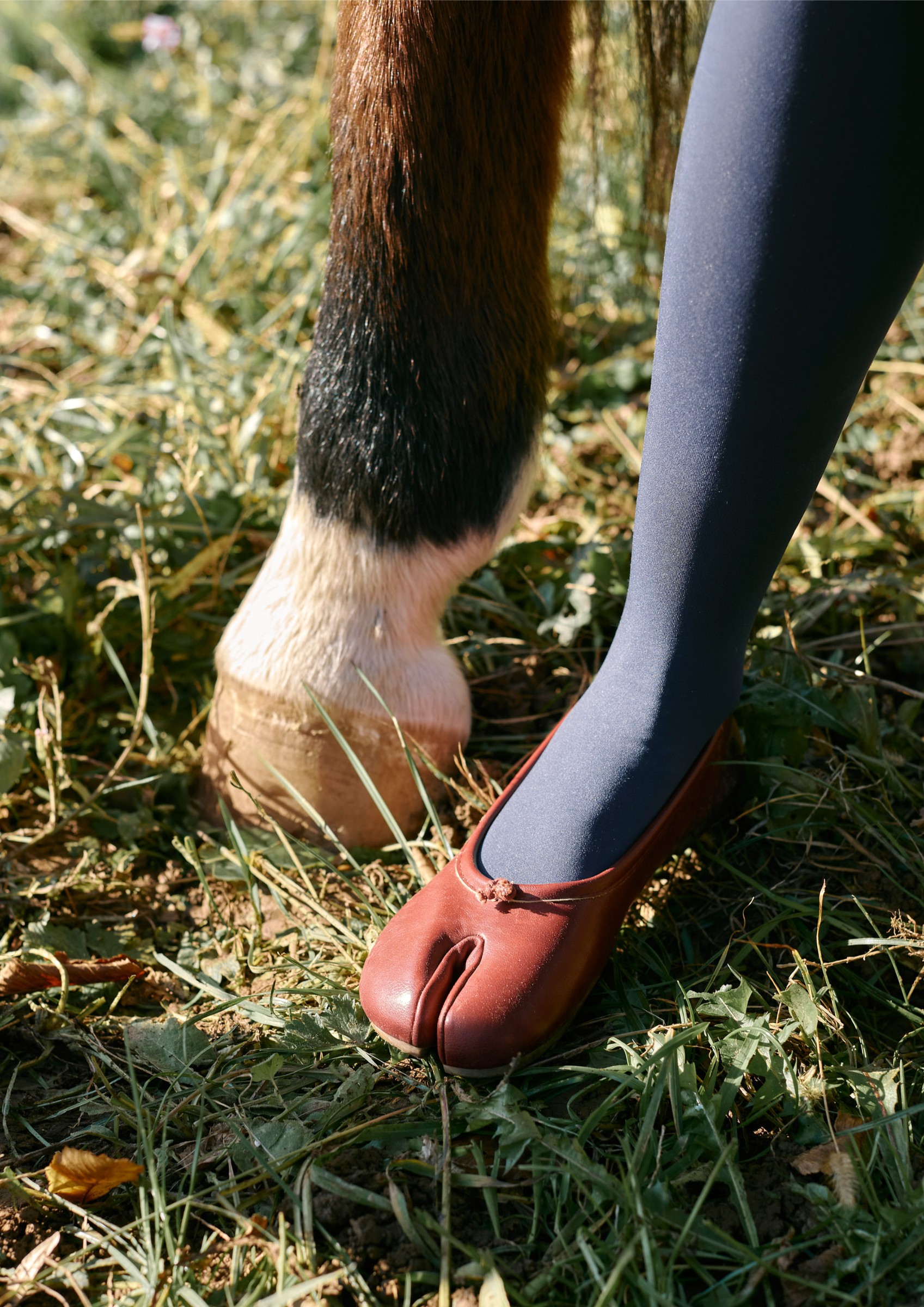 Red split-toe shoe beside horse hoof on grassy ground.