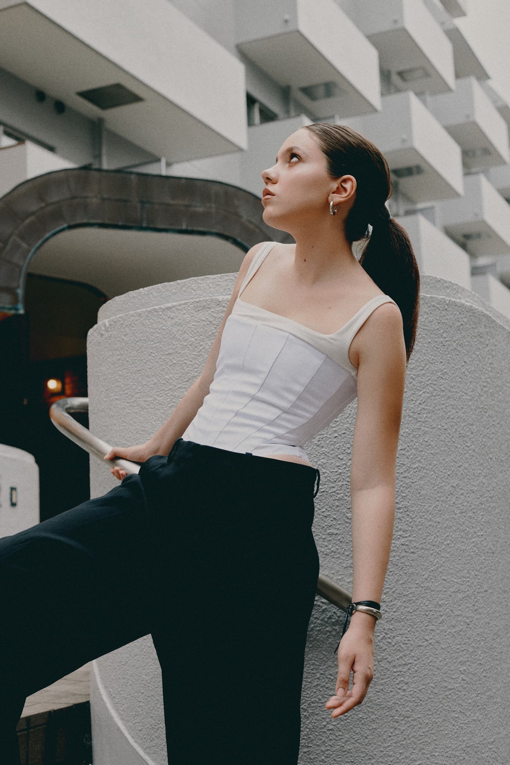 Person looking up in modern architecture setting, wearing stylish white top and black pants.