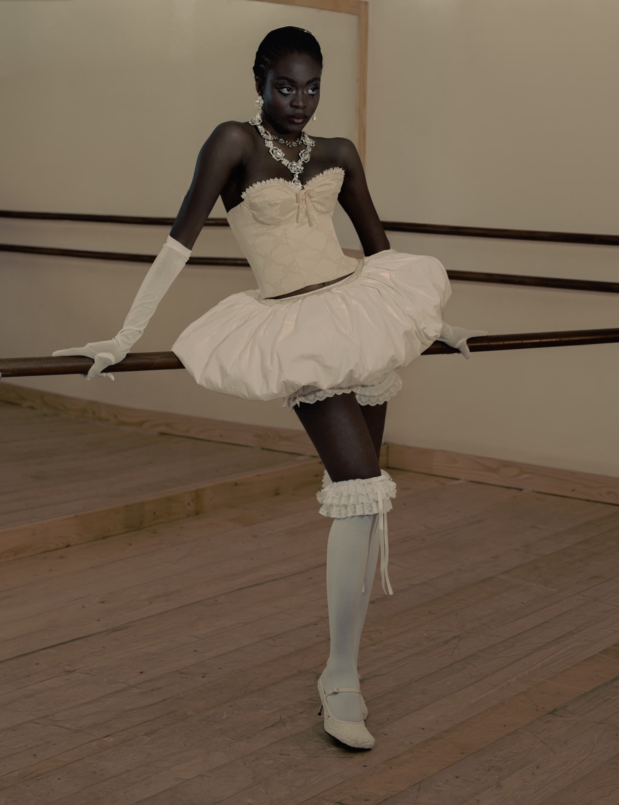 Ballet dancer in elegant white outfit leans gracefully on a barre in a dance studio.