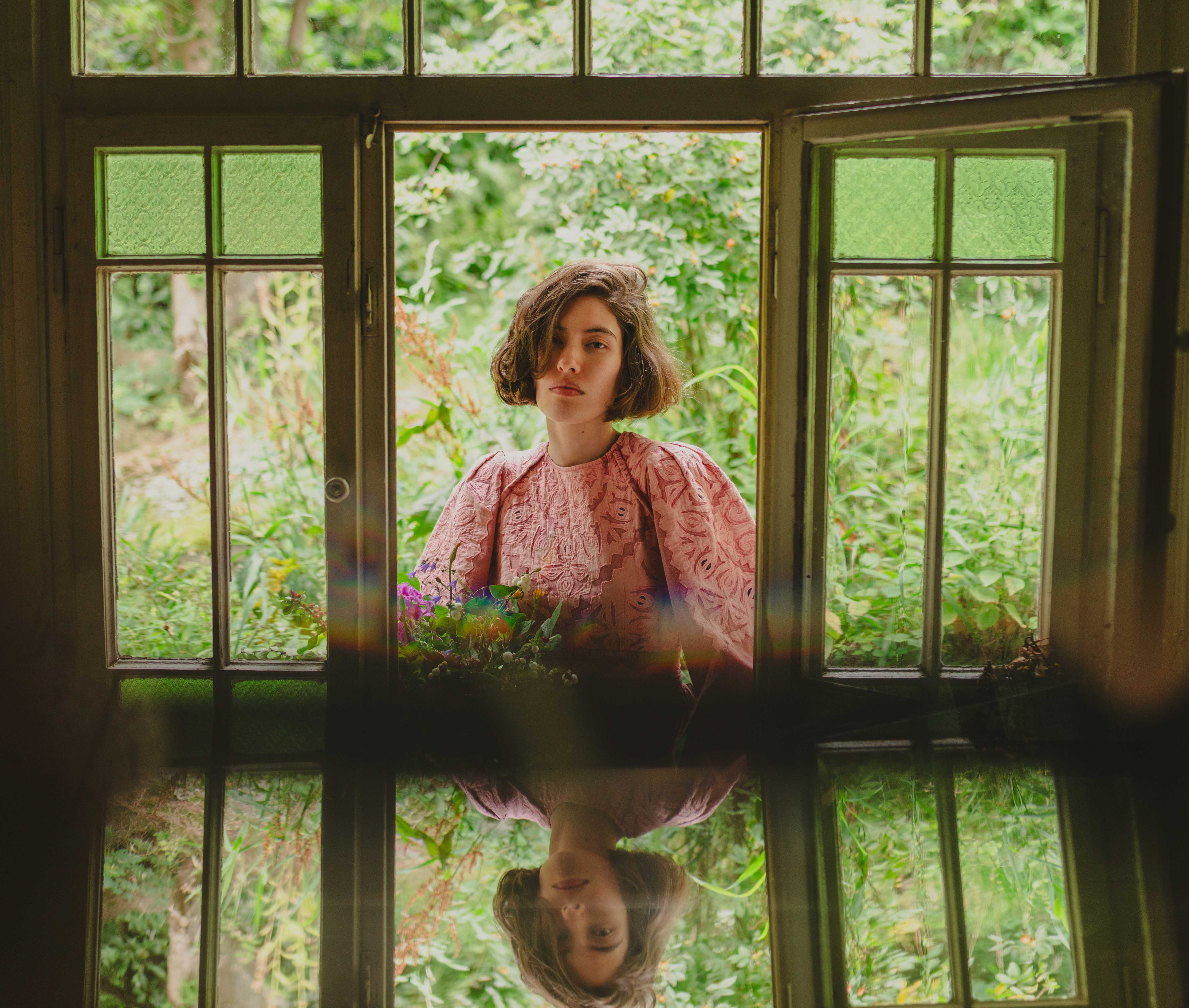 Woman in pink dress at a window with greenery background and reflection.