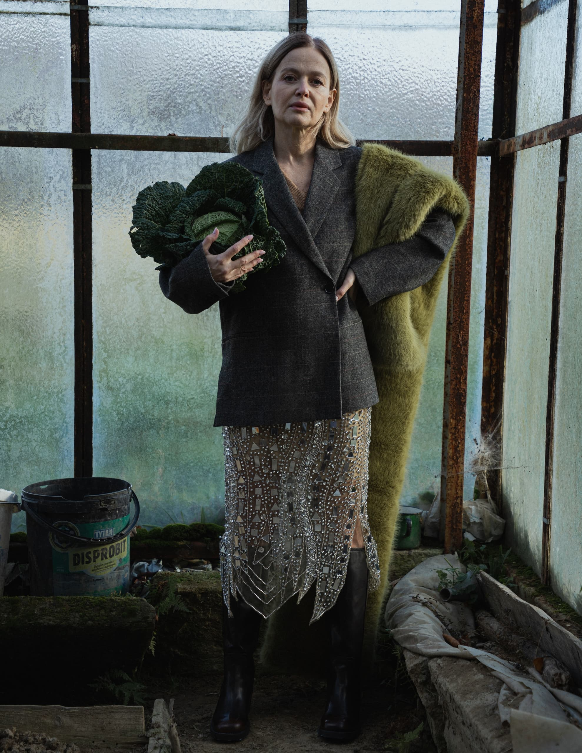 Fashionable woman in chic coat and glittering skirt holding cabbage in greenhouse.