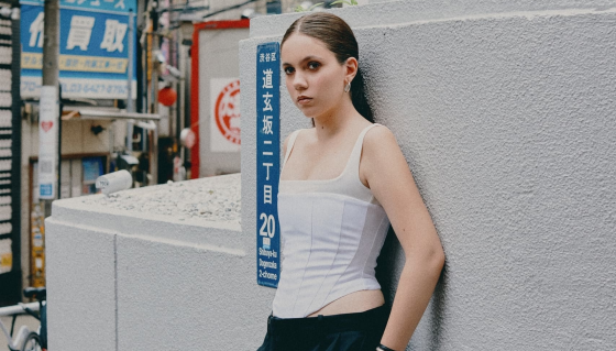 Young woman in a white top leans on a textured wall in an urban setting.