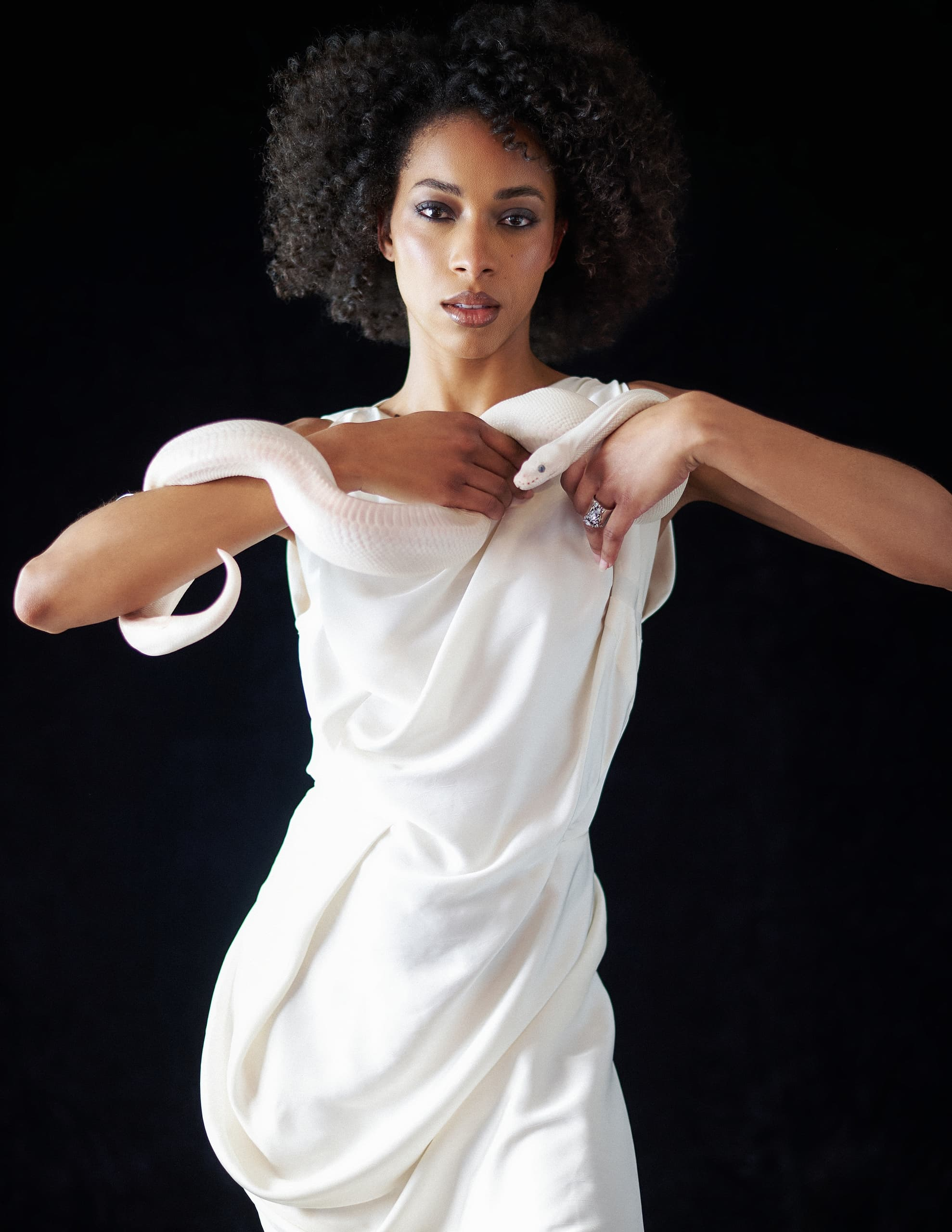 Woman in white dress holding a white snake against a dark background.