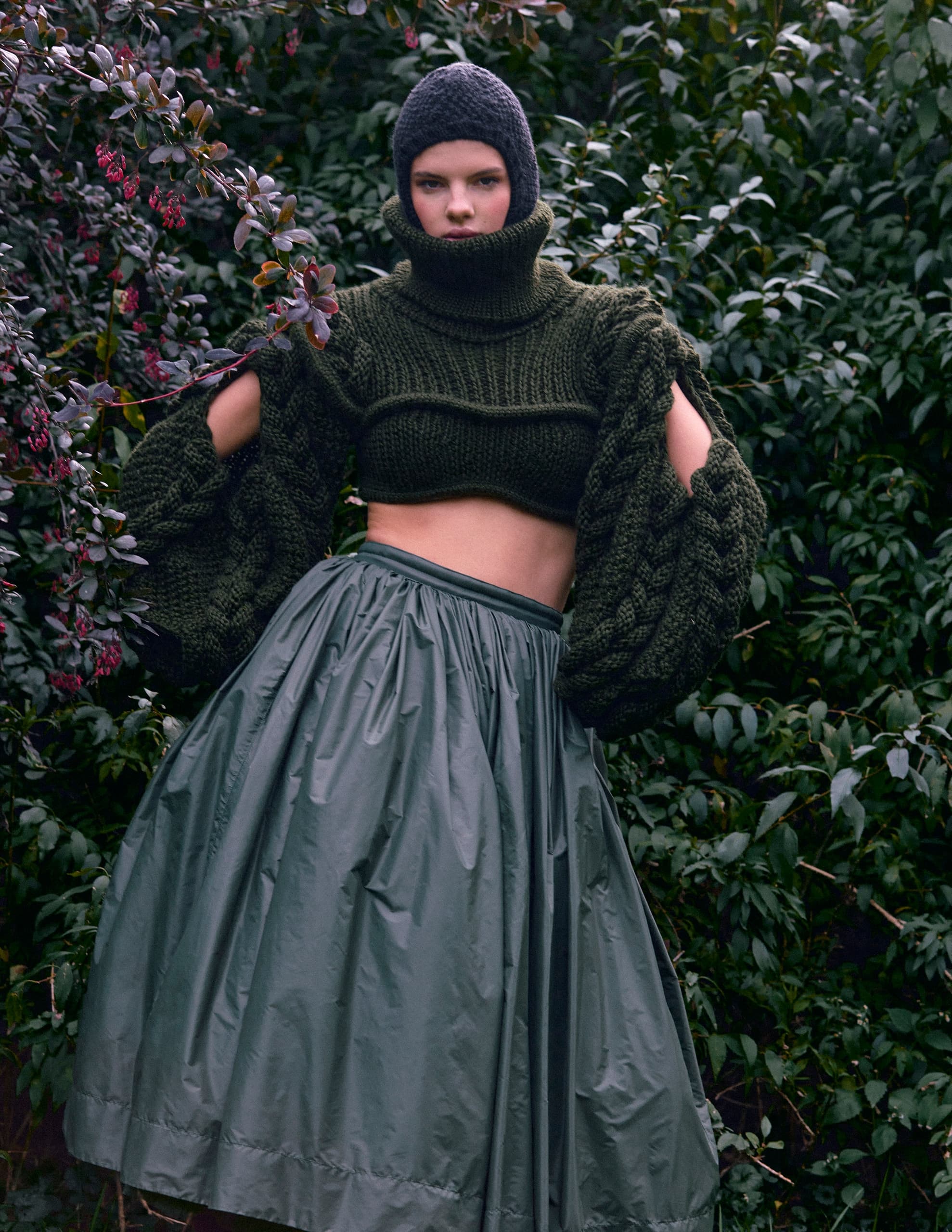 Fashion model in a green knit outfit and skirt in a lush garden setting.