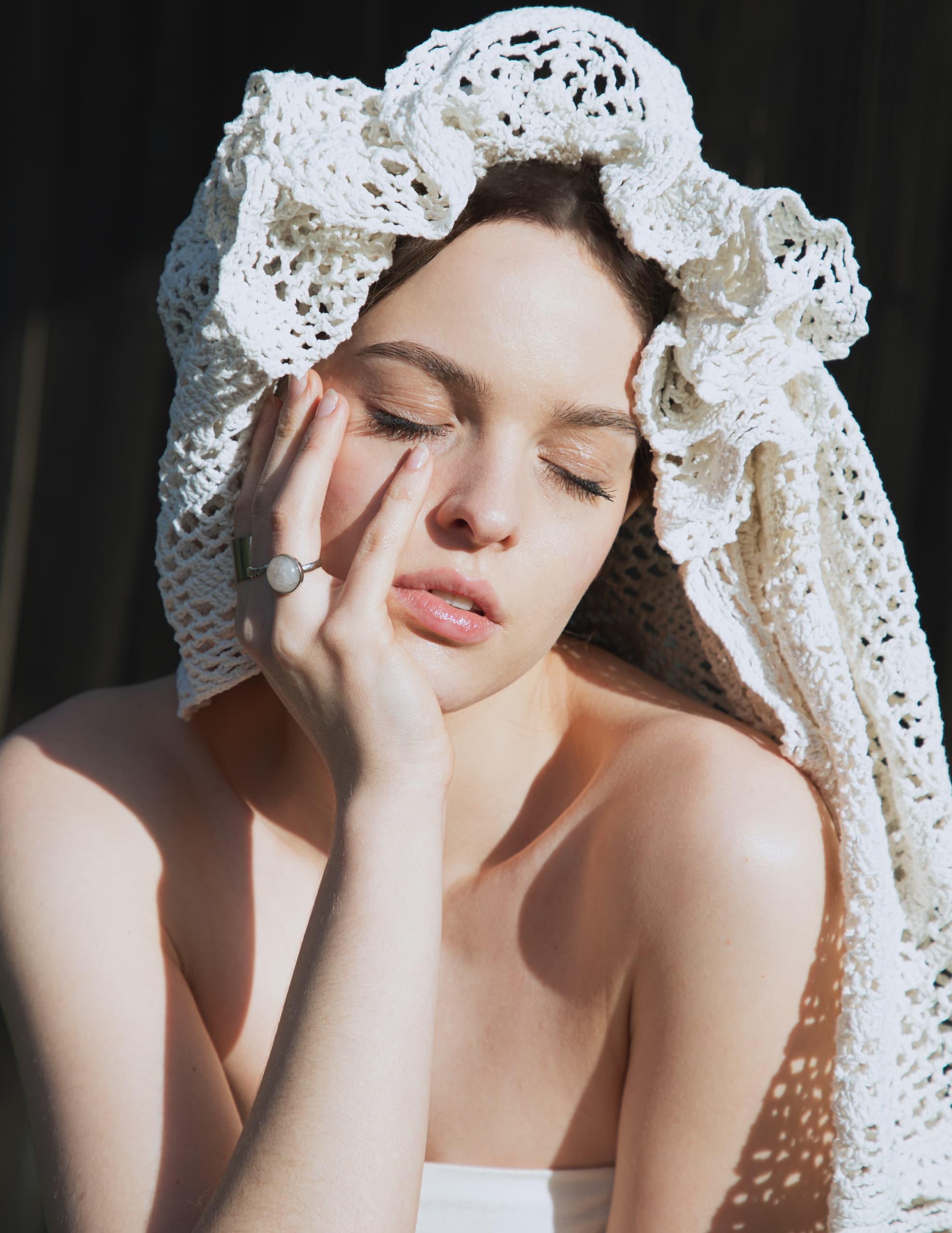 Person with eyes closed, wearing a white crochet headpiece in sunlight.