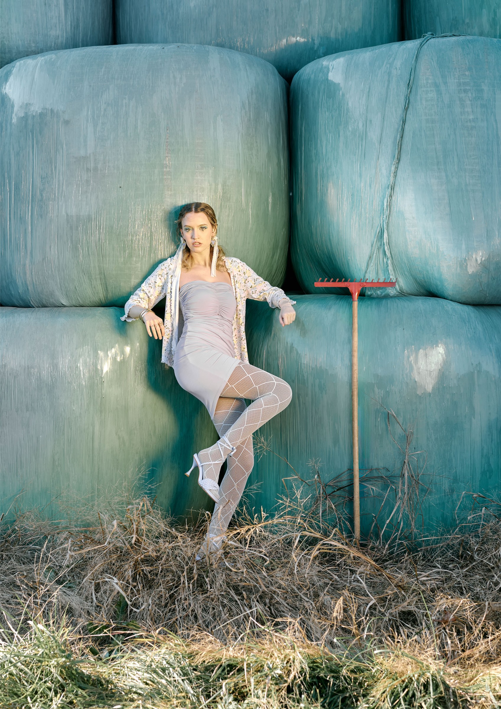 Fashionable woman in stylish outfit poses by large hay bales and a red rake.