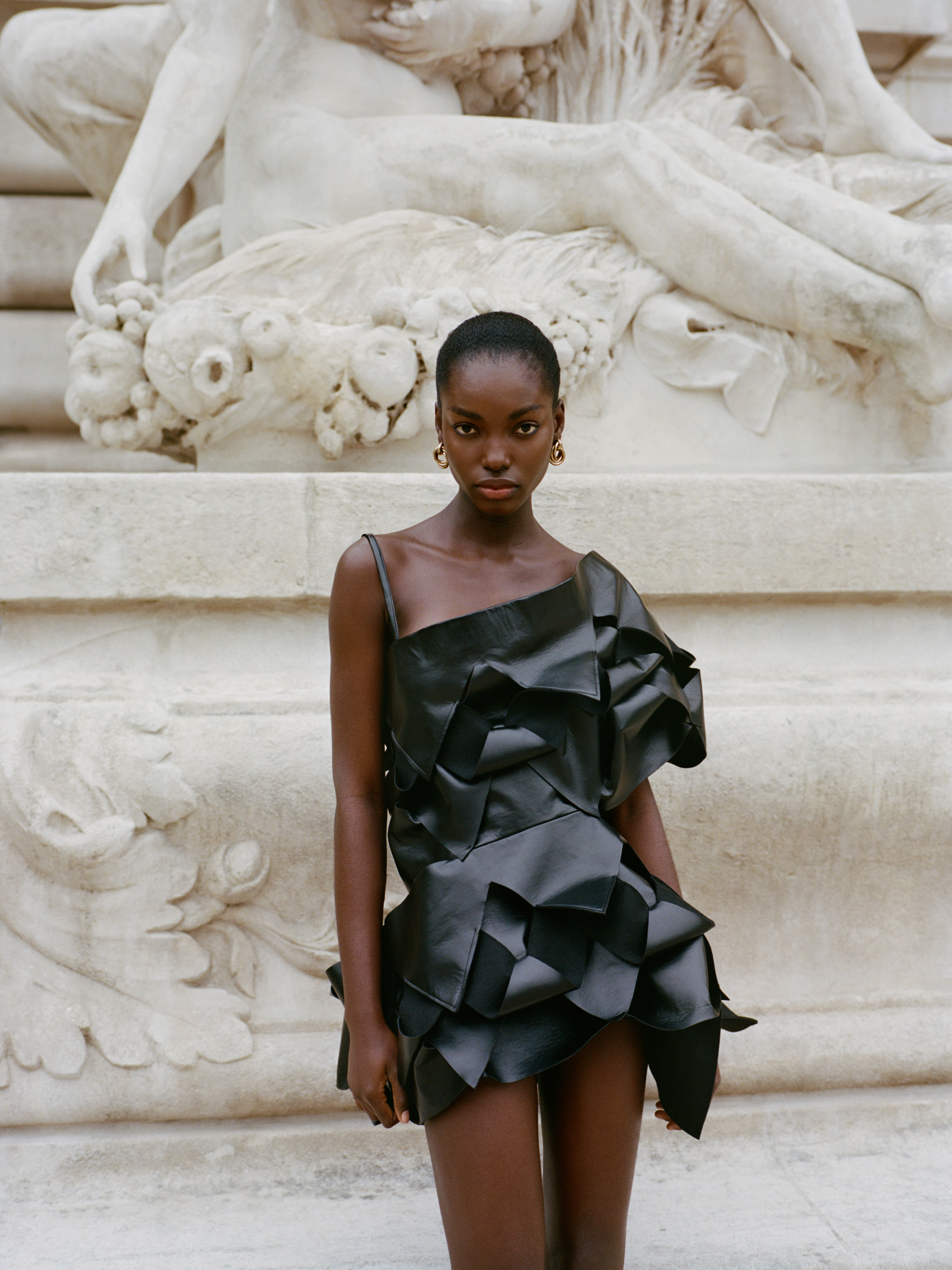 Model in black fashion dress posing by intricate stone sculpture.